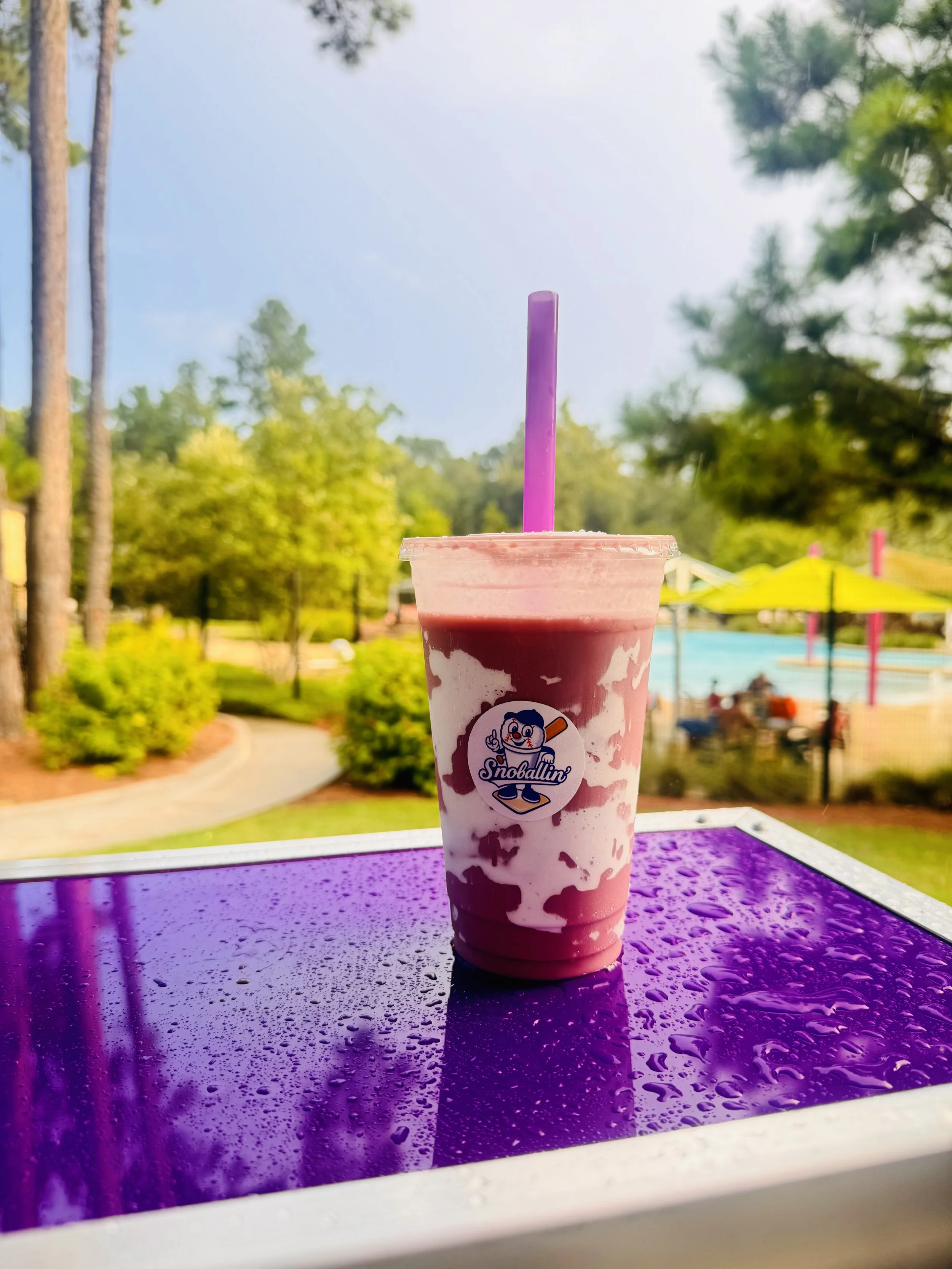 A pink and white ice blended drink with a purple straw on a purple reflective surface outdoors, with trees and a swimming pool in the background.