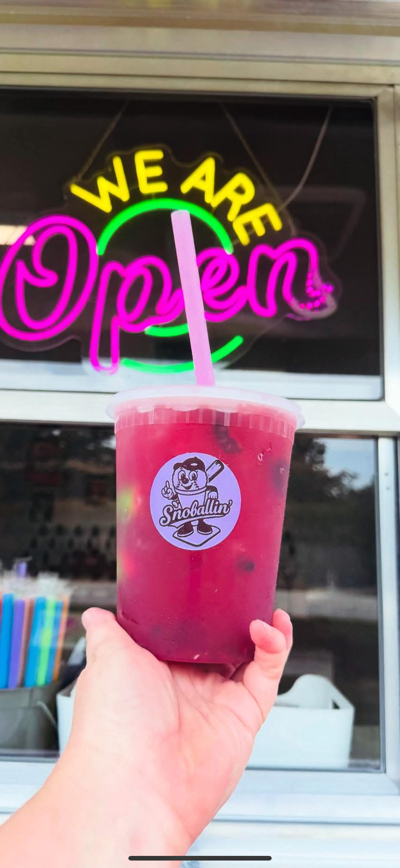 A hand holding a pink drink with a straw in front of a neon sign that says 'We Are Open'.