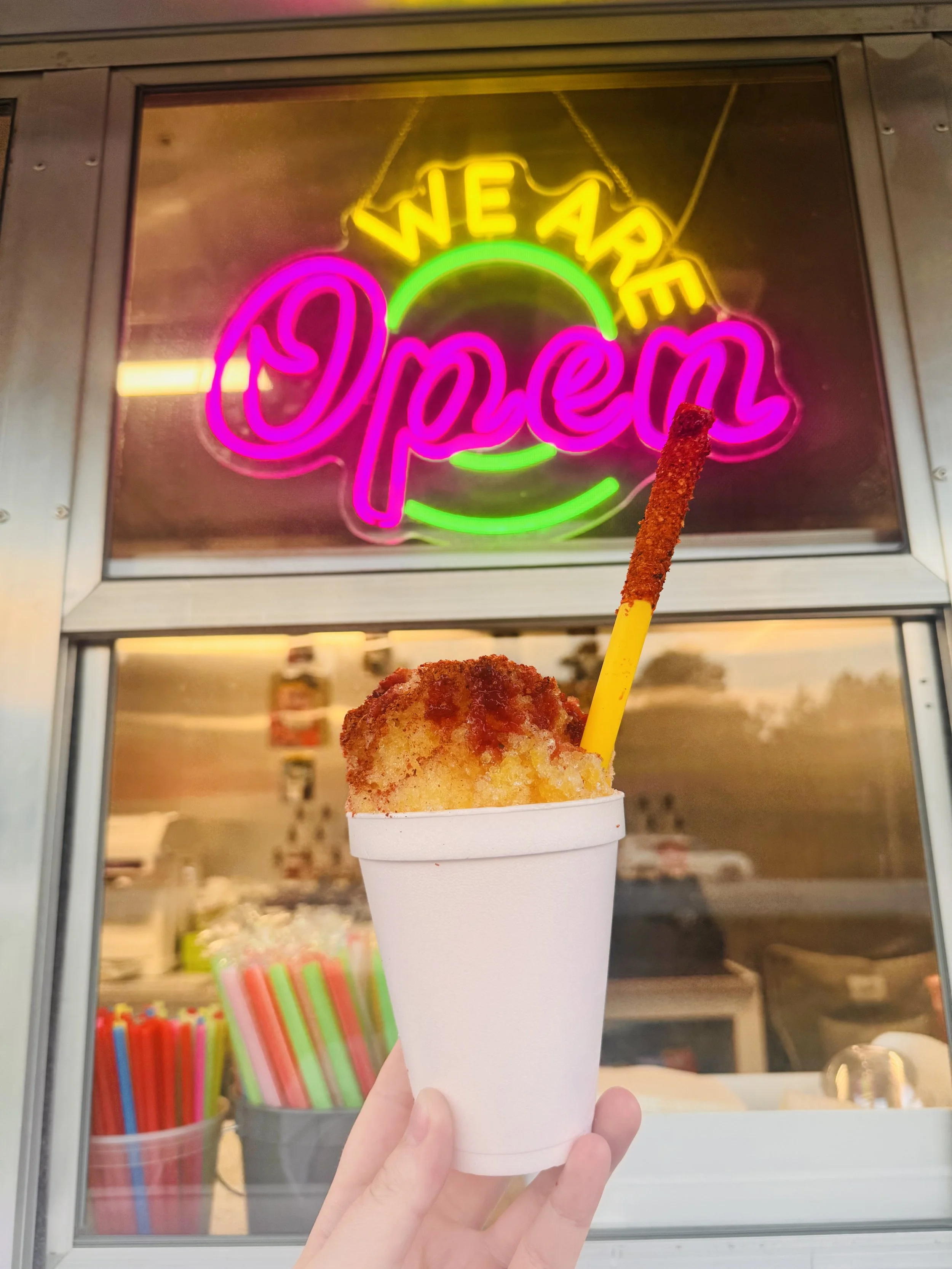 A hand holding mangonada, in front of a neon sign that says 'WE ARE OPEN' in pink, yellow, and green lights.