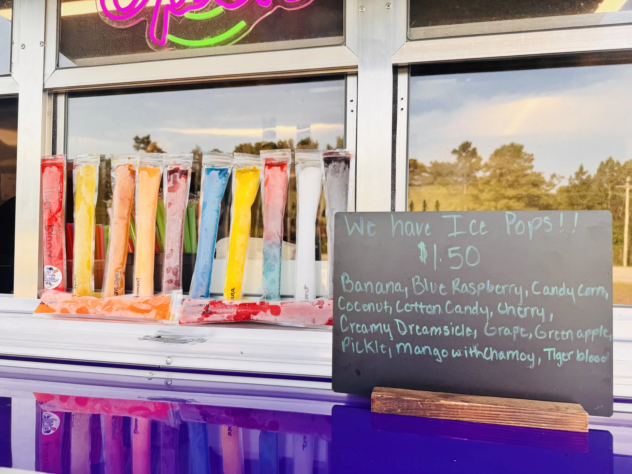 A food truck window displaying colorful ice pops in clear plastic bags and a chalkboard sign listing available flavors and prices.