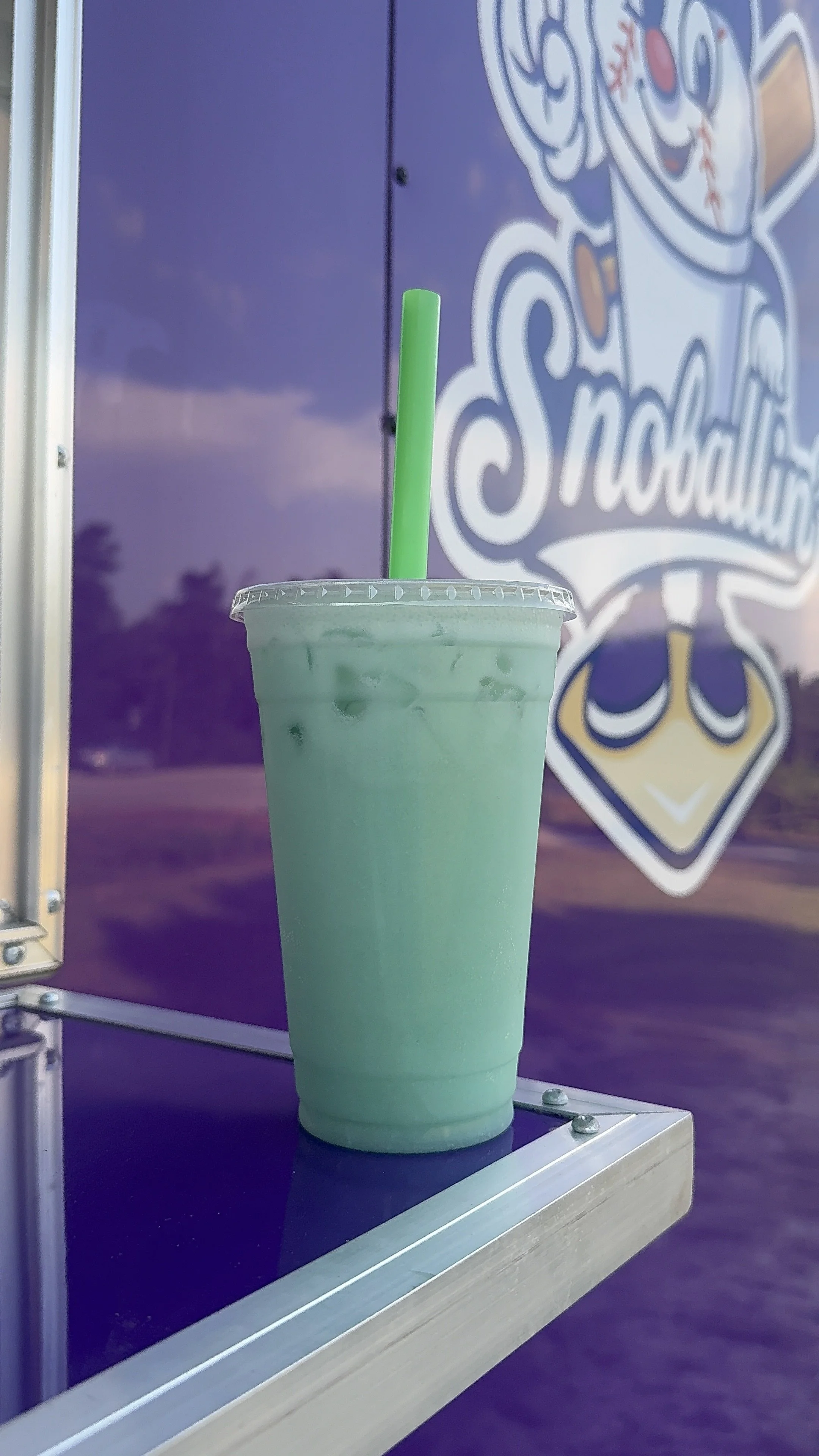 A light green milkshake with ice in a clear plastic cup, topped with a clear plastic lid and a green straw, sitting on a metal surface in front of a purple backdrop with the word 'Snowball' partially visible.