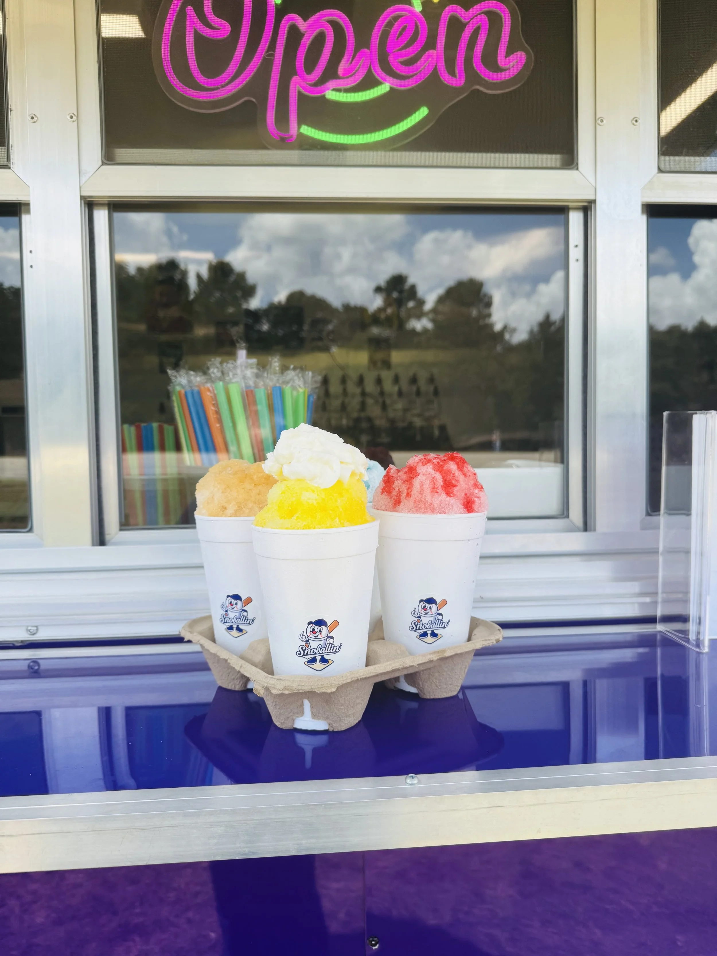 Four cups of colorful shaved ice topped with whipped cream and flavored syrup, placed on a tray at a snow cone stand with an 'Open' sign in neon above.