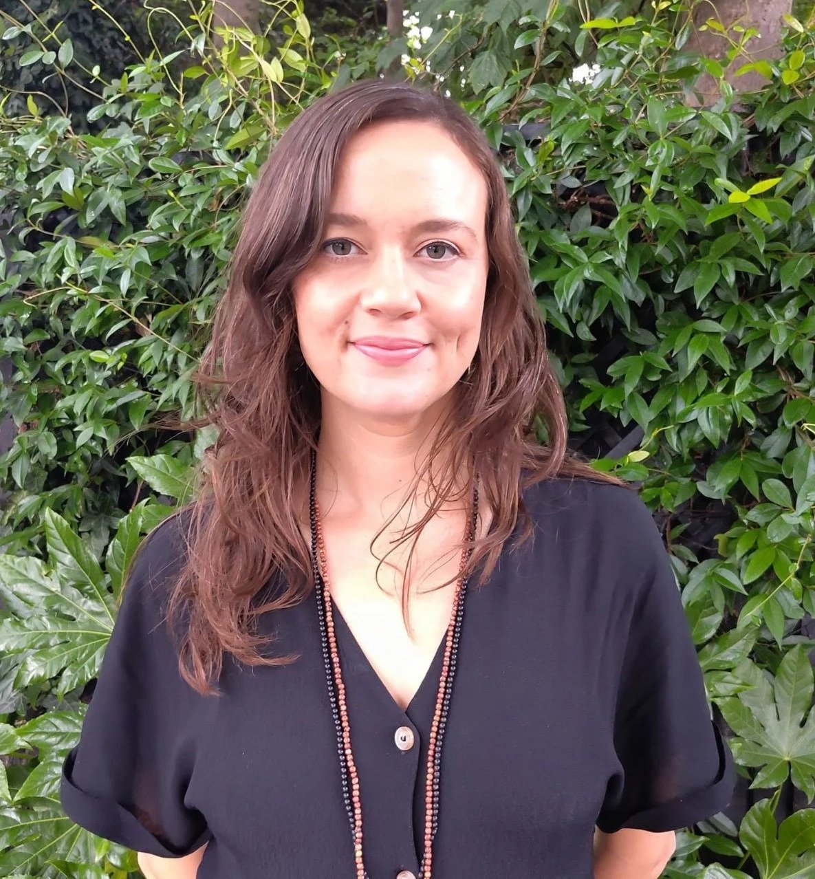 A photo of Ellen Amorina. She has wavy brown hair and fair skin smiling in front of green leafy bushes, wearing a black top and a beaded necklace.