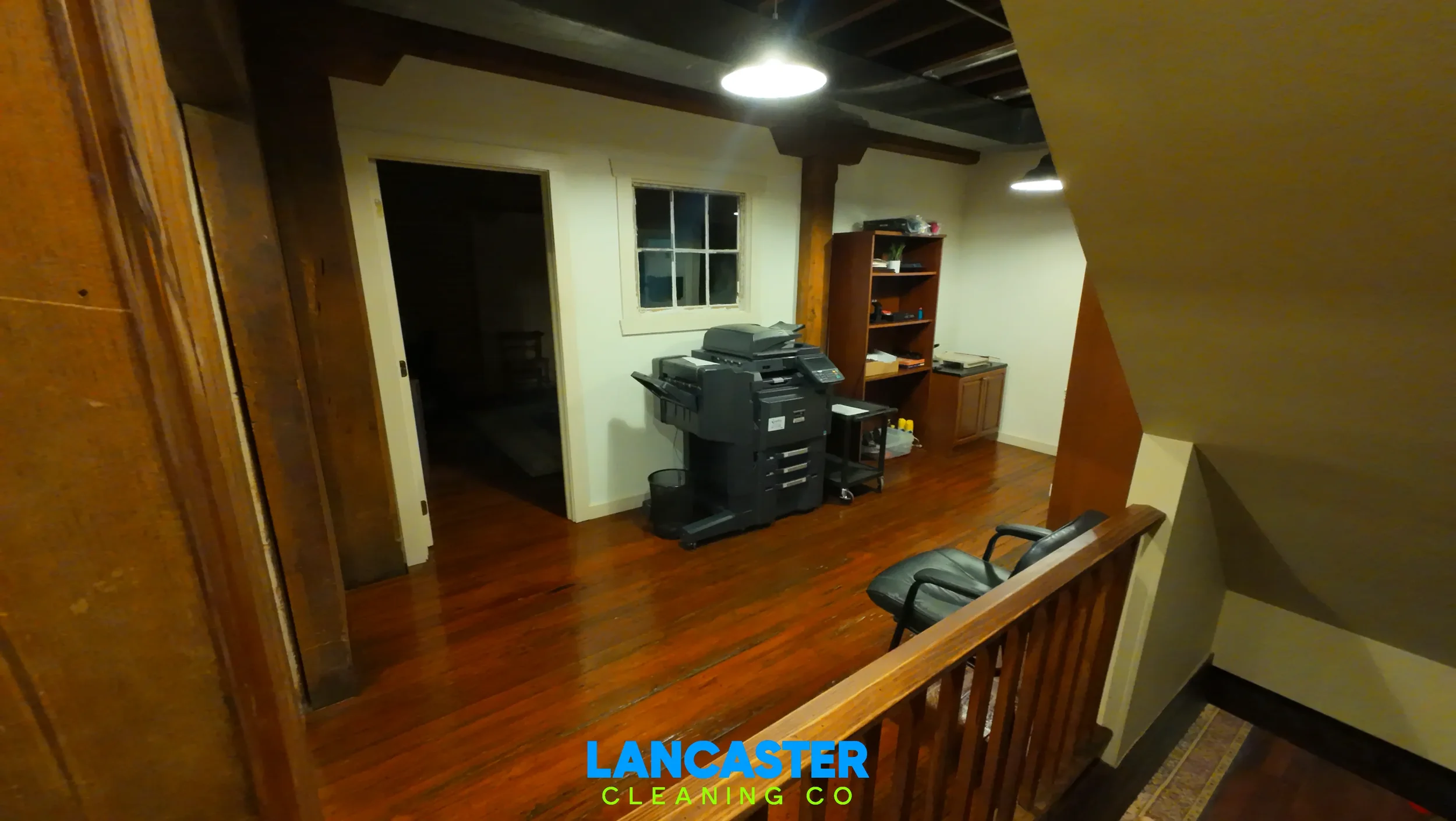 Office cleaning in Lancaster, PA featuring polished hardwood floors, exposed wooden beams, a multifunction printer, shelving unit, and stair railing, completed by Lancaster Cleaning Co.