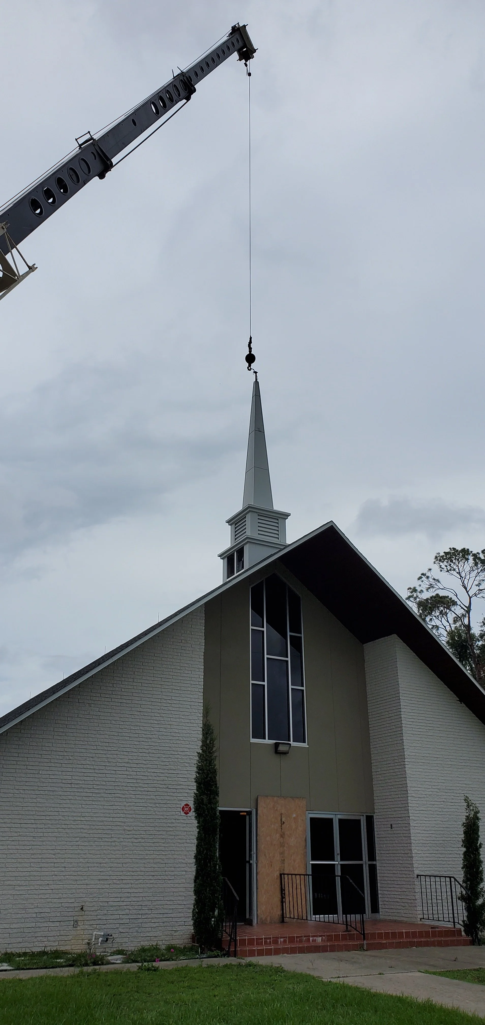 Construction crane lifting church steeple onto a building under an overcast sky.