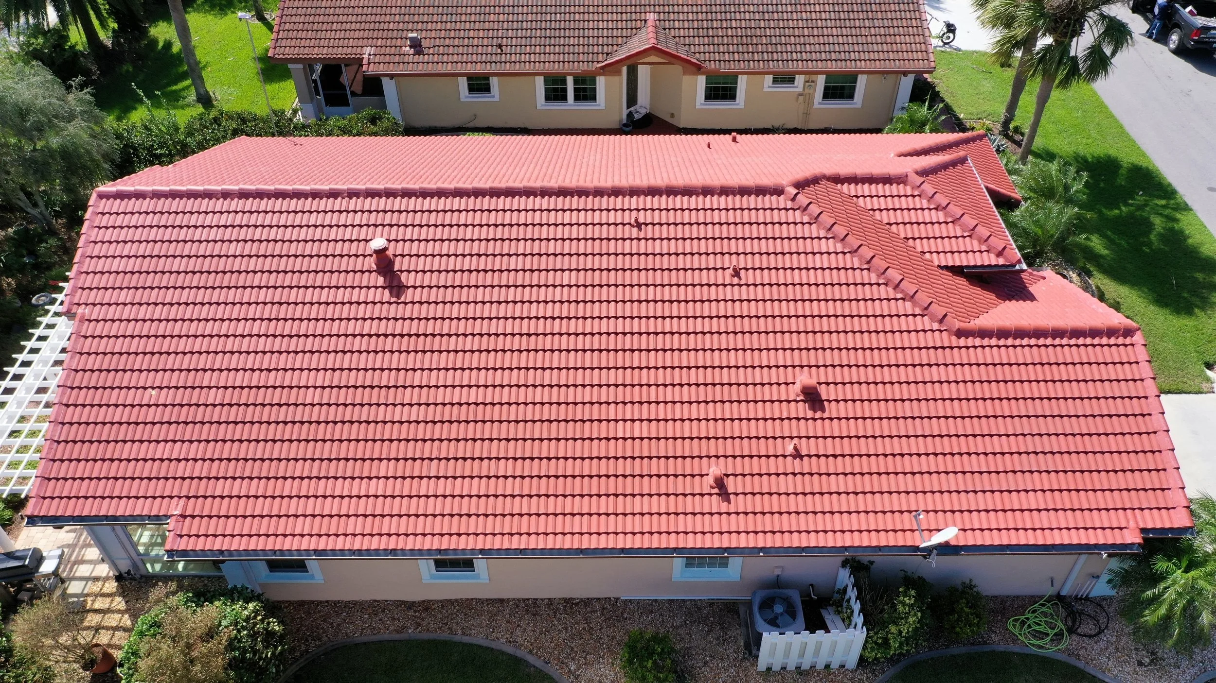Aerial view of a house with a red tiled roof, surrounded by green lawn, trees, and neighboring houses.