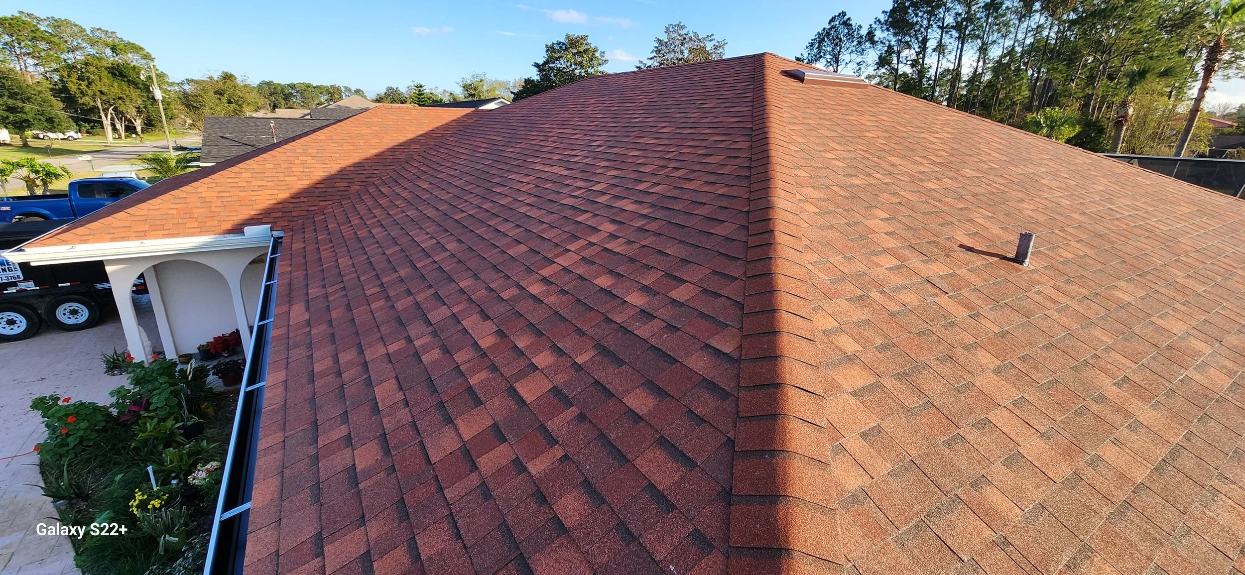 Aerial view of a house roof with red asphalt shingles, a vent pipe, and a yard with plants, flowers, and a parked black truck with a trailer.