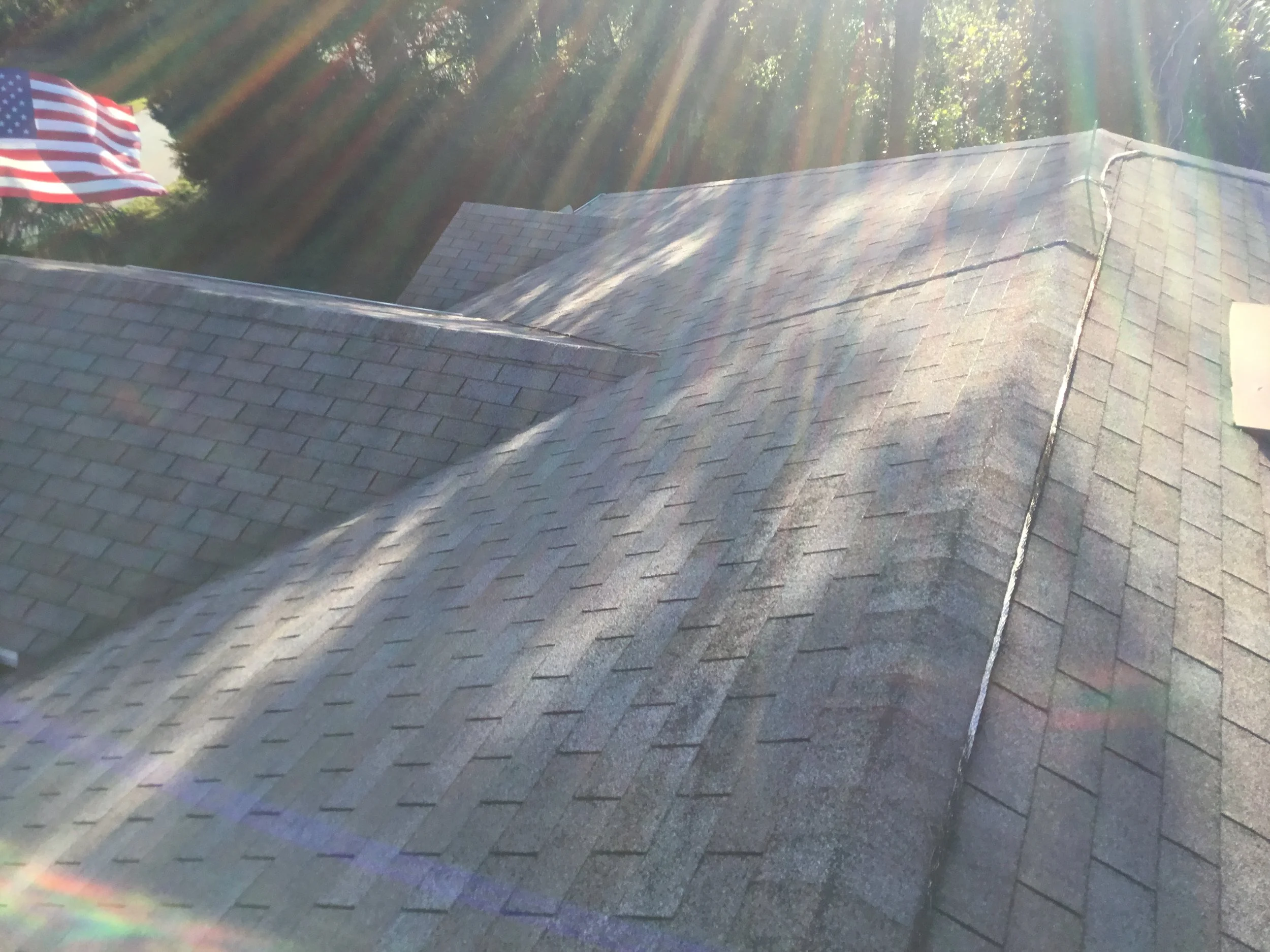 Close-up view of a roof with asphalt shingles, sunlight creating lens flare, and American flag partially visible in the background.
