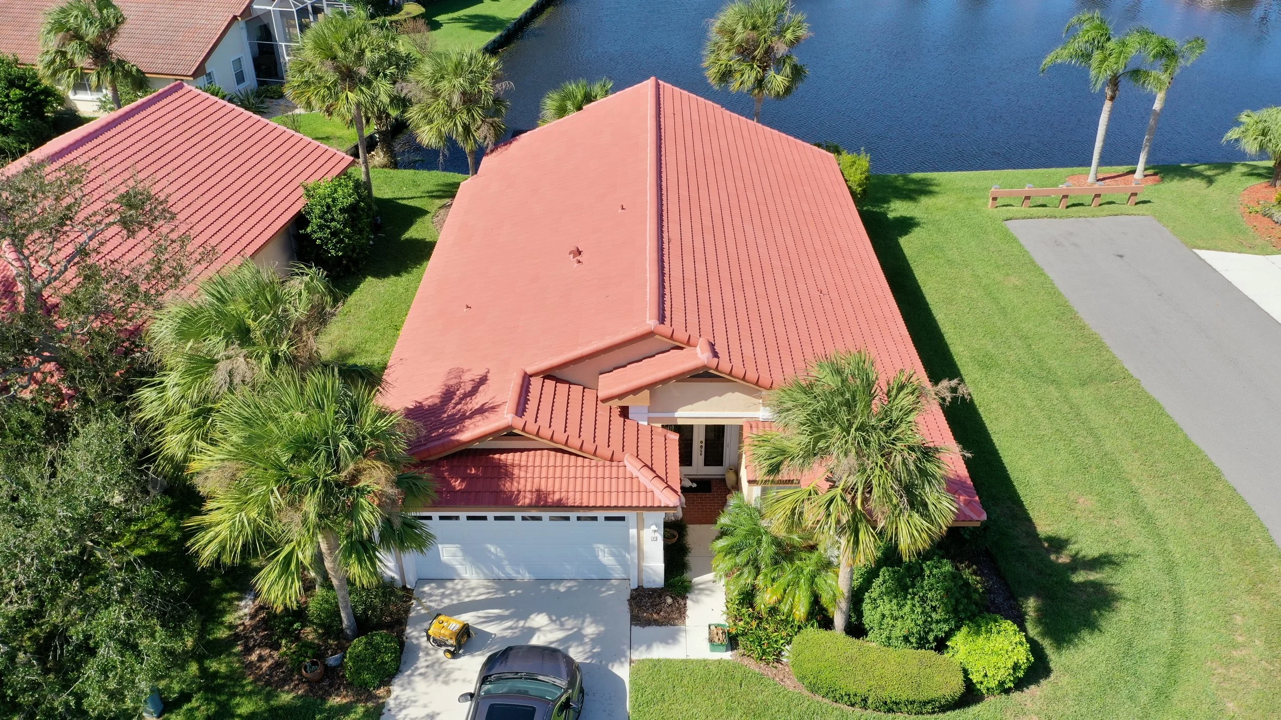Aerial view of a house with a red tile roof, surrounded by green grass and palm trees, next to a water body, with a driveway and parked cars.