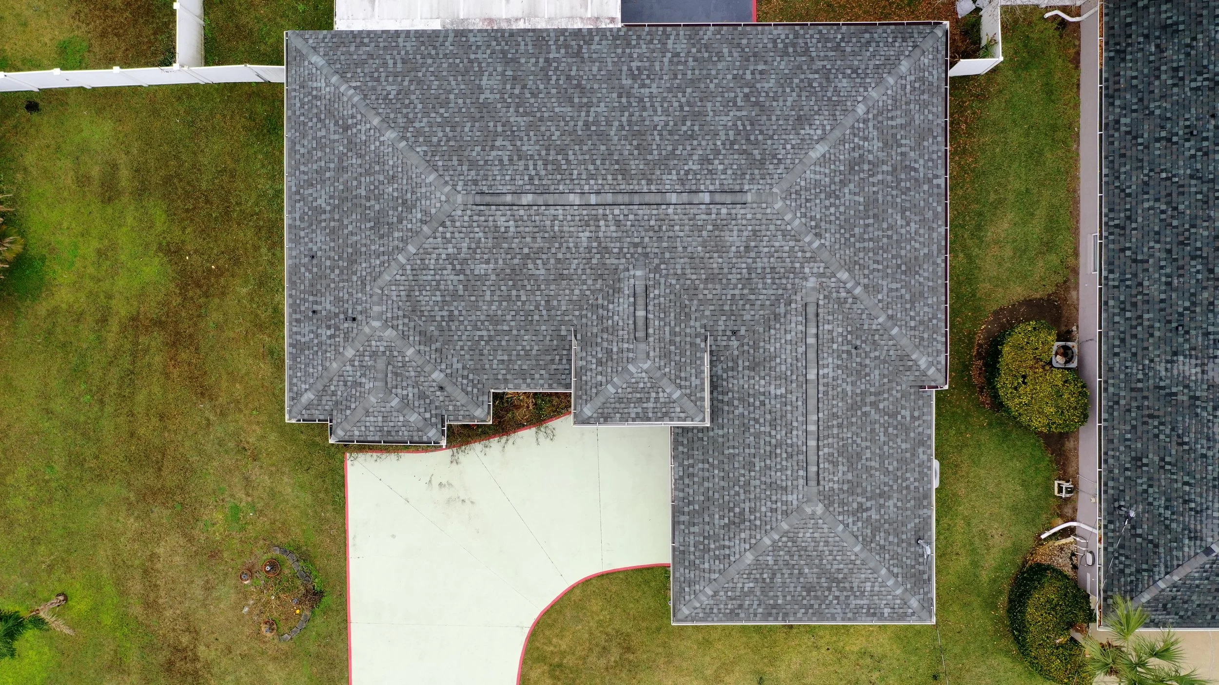 Aerial view of a house with a dark gray shingle roof, surrounded by a fenced backyard with grass, a curved concrete driveway, and some plants and trees.