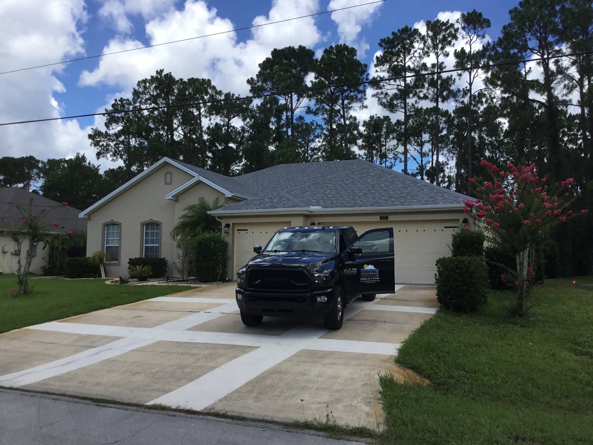 A black pickup truck parked in front of a beige house with a two-car garage, surrounded by green grass, small trees, bushes, and flowering plants, with a cloudy blue sky and tall trees in the background.