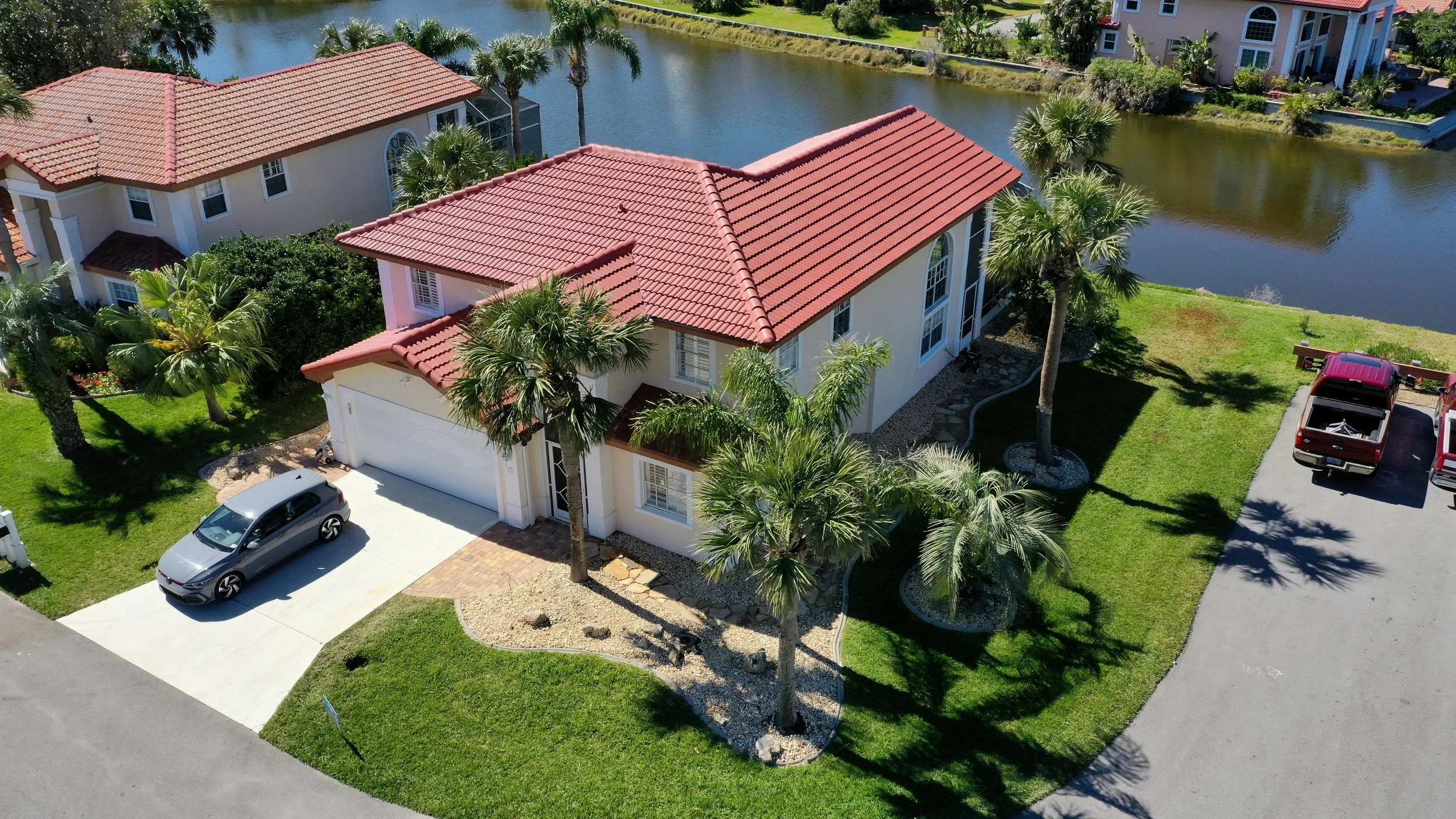 Aerial view of a two-story house with a red tile roof, surrounded by palm trees and green lawns, next to a water body, with a driveway, several parked cars, and neighboring houses in the background.