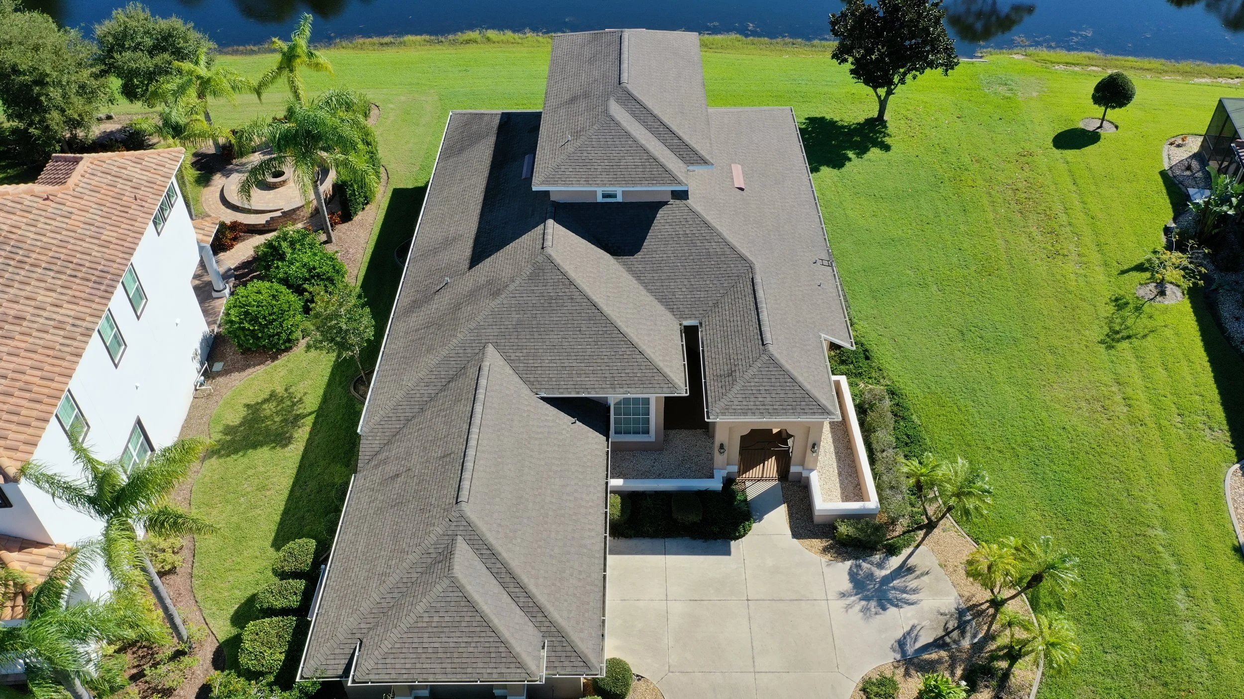 Aerial view of a house with a dark gray roof, surrounded by green lawns, trees, and neighboring houses, with a body of water in the background.