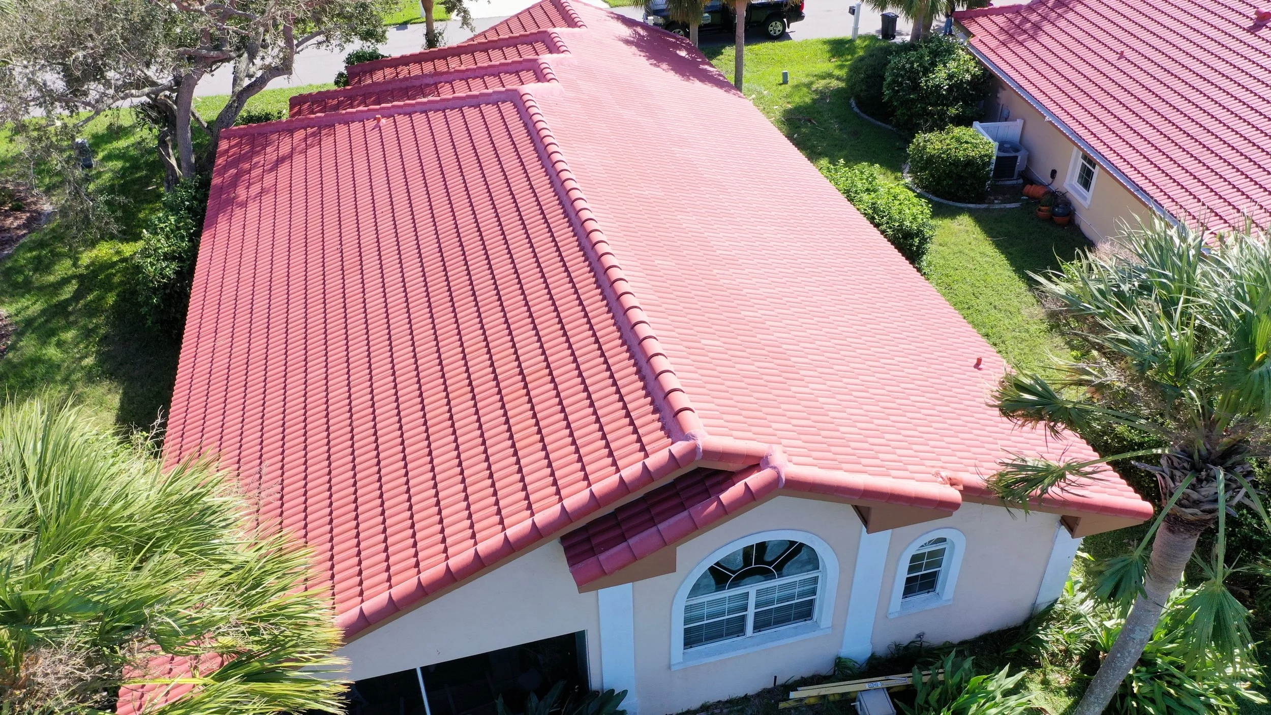 Aerial view of a house with a red tile roof, white exterior walls, surrounded by green trees and bushes, with a small yard and garden area.