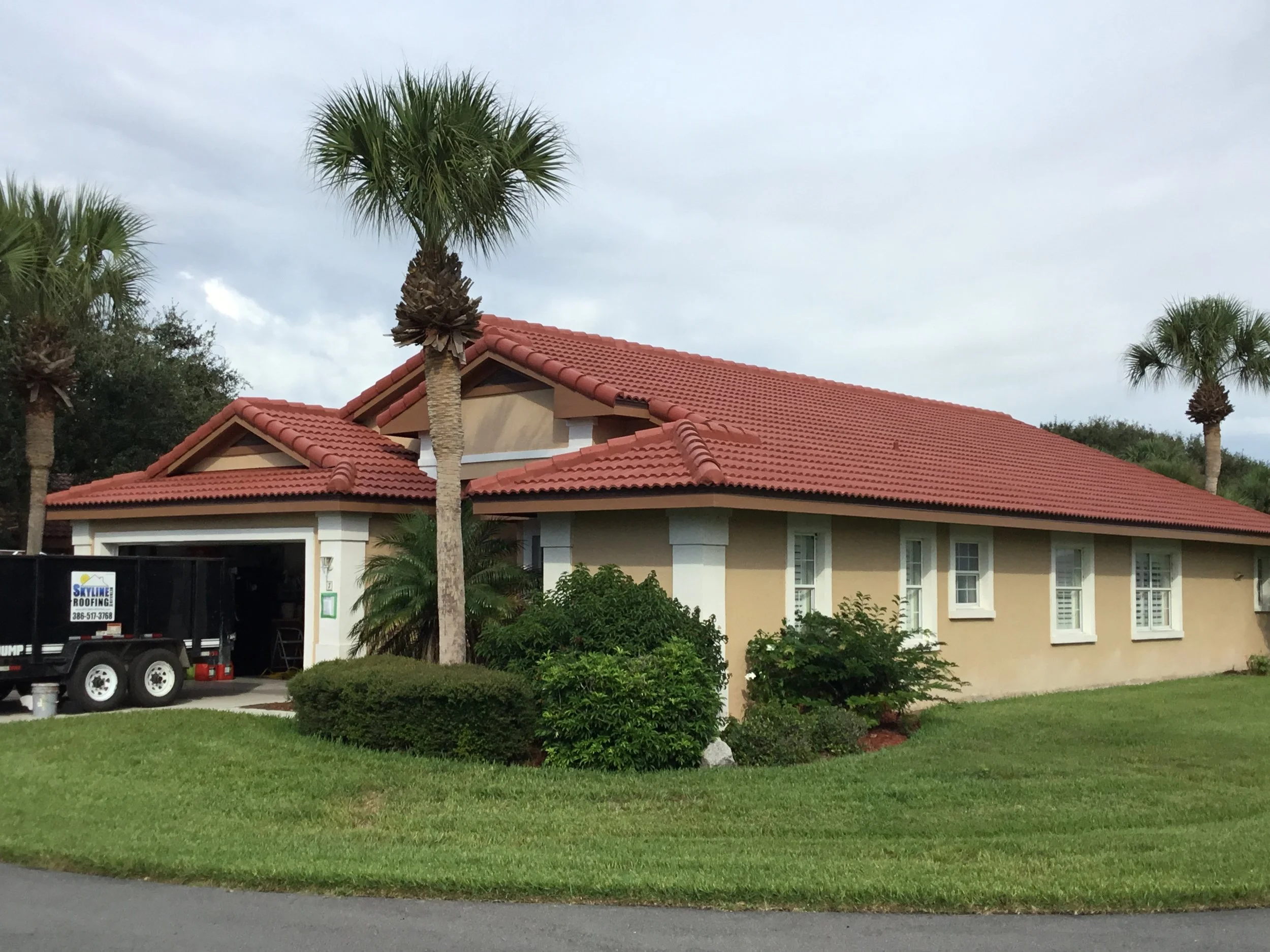 A house with a red-tiled roof, beige walls, and white trim, surrounded by palm trees and green bushes, on a cloudy day.