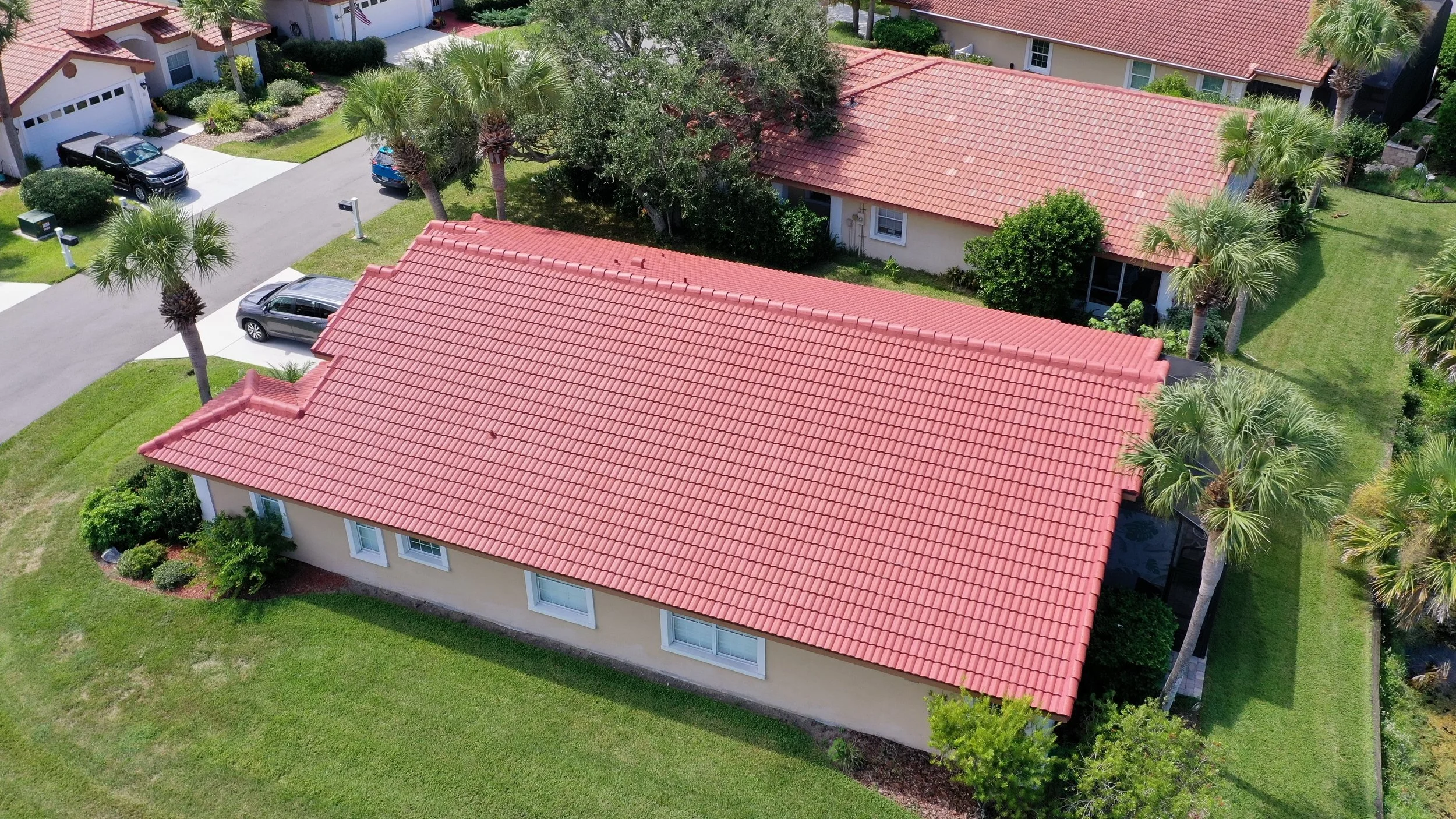 Aerial view of a residential neighborhood with houses featuring red-tiled roofs, surrounded by well-maintained green lawns, trees, and parked cars.
