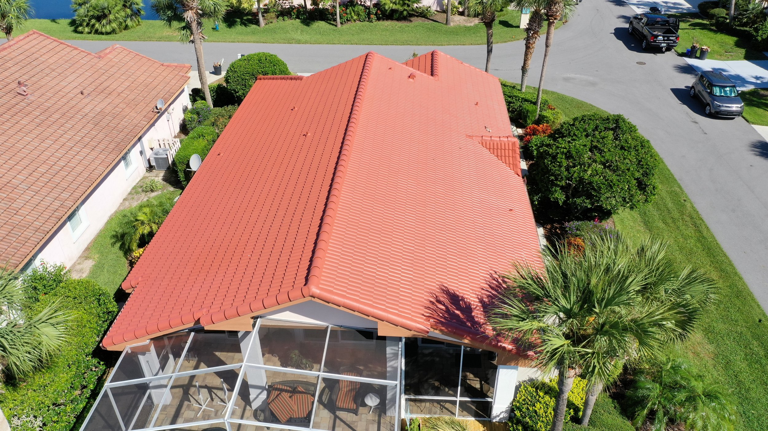 Aerial view of a house with a red tile roof, surrounded by green trees, bushes, and a lawn, with parked cars on the street.