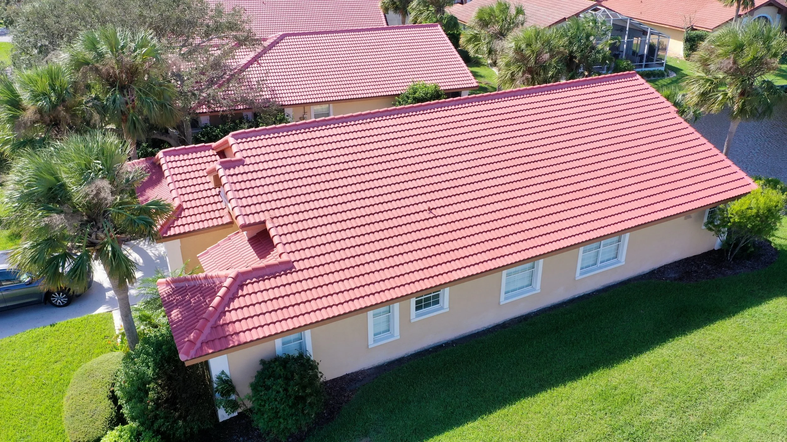 Aerial view of a house with a red tile roof, surrounded by green grass, palm trees, and neighboring houses with similar roofs.