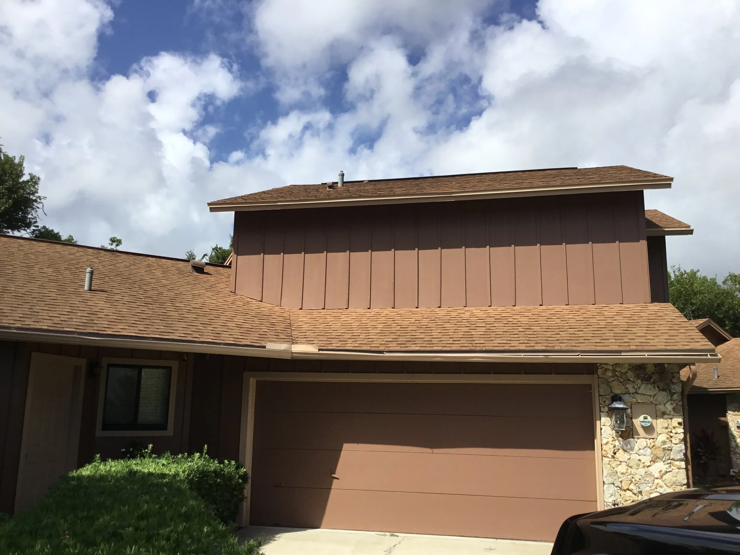 A two-story house with a brown garage door, brown siding, stone accents, a brown shingle roof, a small window to the left of the garage, and a bush in front, under a partly cloudy sky.