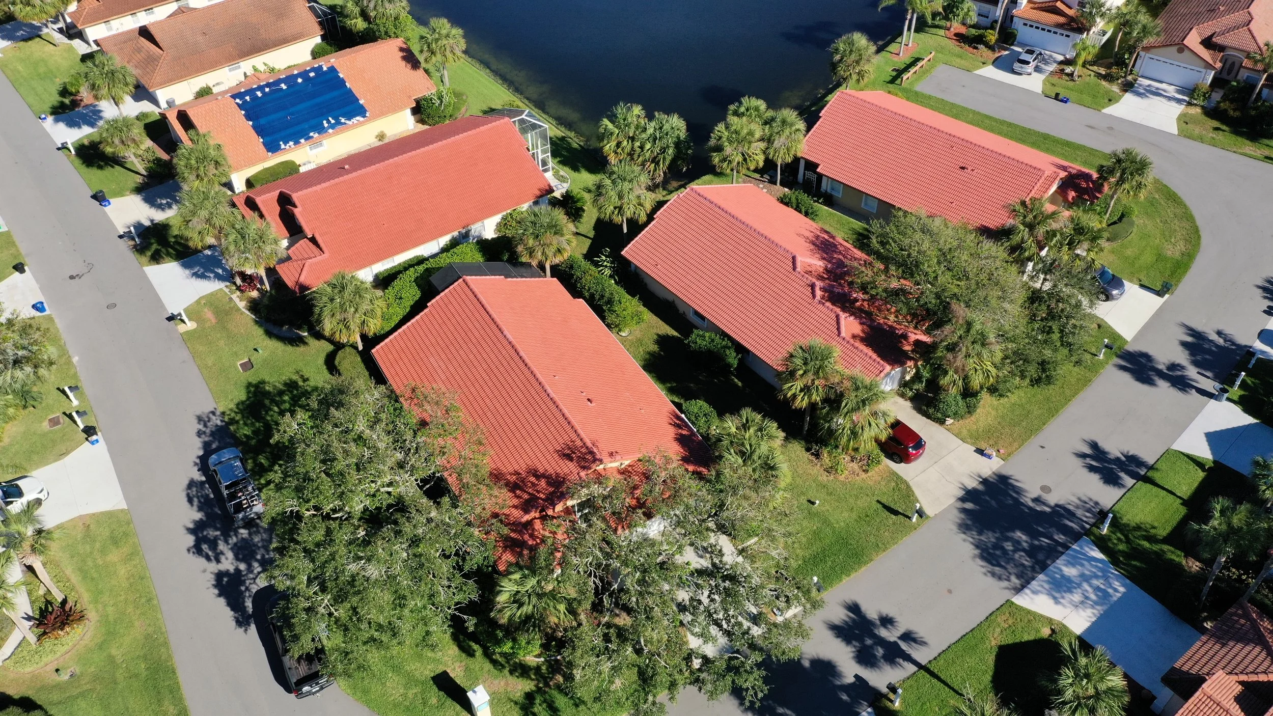 Aerial view of a residential neighborhood with houses having red-tiled roofs, green lawns, palm trees, cars parked in driveways, and a water body in the background.