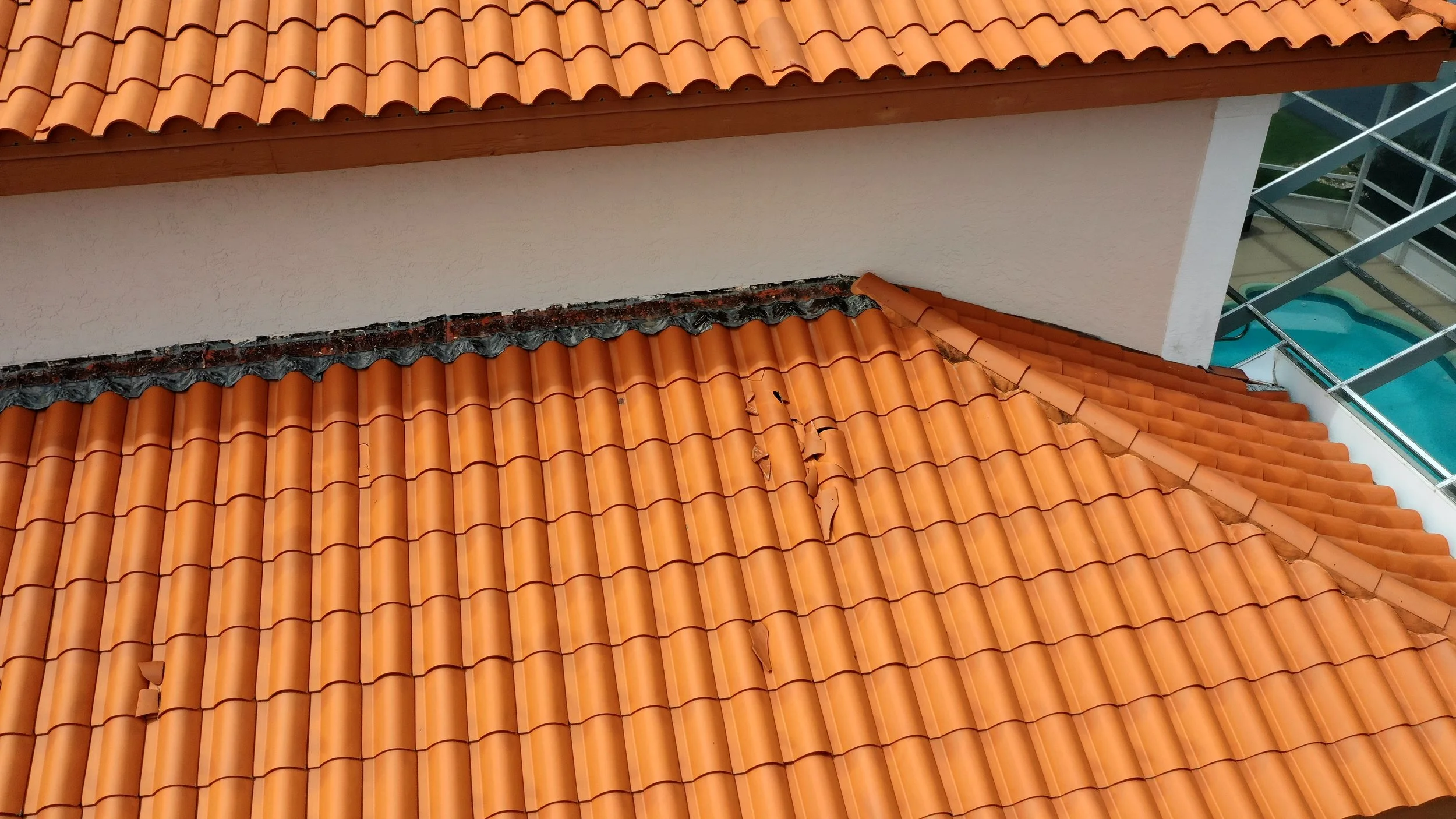 Clay tile roof showing both damaged tiles and failing wall flashing where the roof meets vertical siding. Cracked and displaced tiles expose underlayment, while missing or improperly layered flashing allows water to track behind the roof system — a c