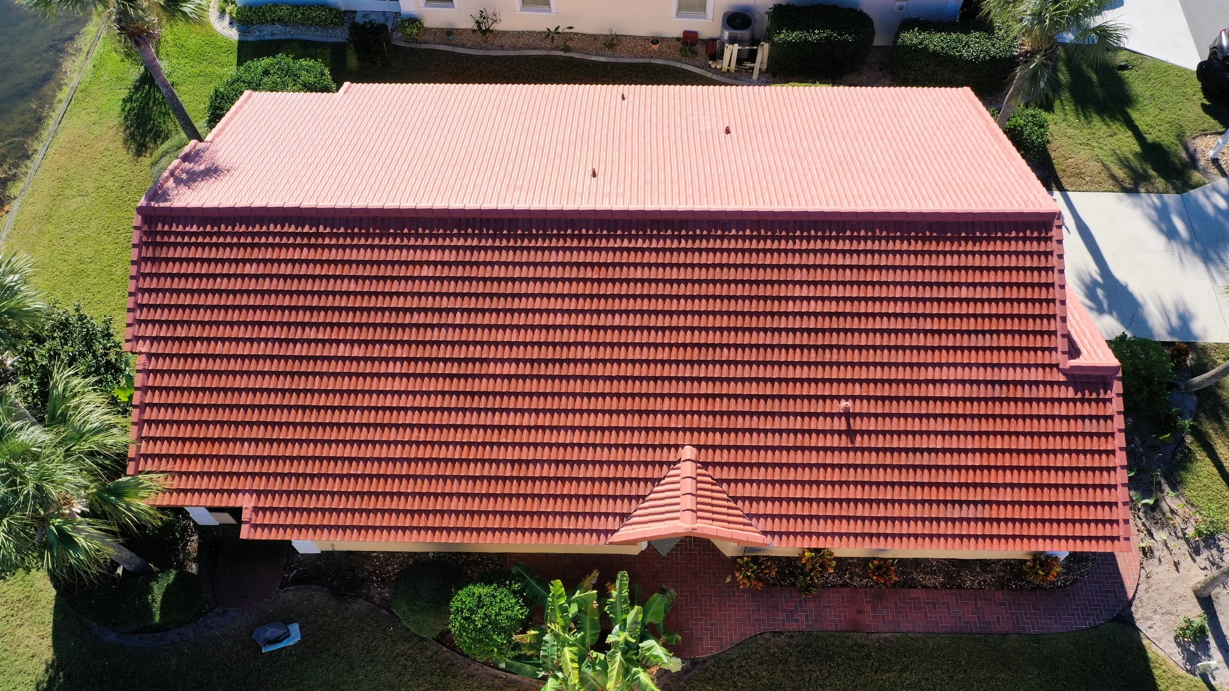 A house with a red tiled roof, surrounded by green lawn, palm trees, and garden shrubs, seen from above.
