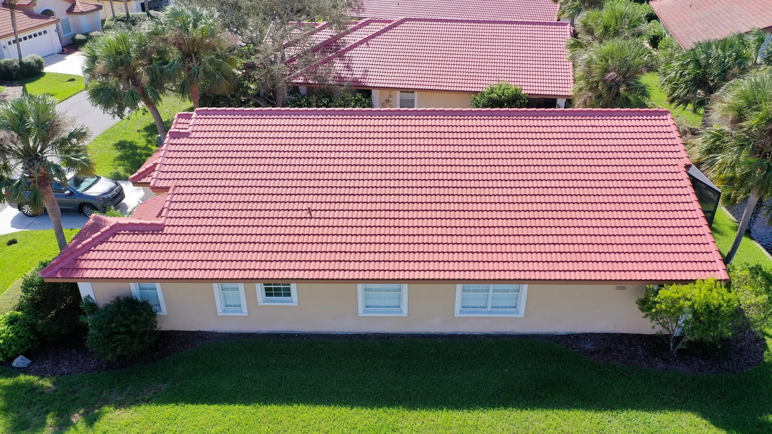 An aerial view of a house with a red tile roof, beige walls, and a well-maintained green lawn, surrounded by palm trees and neighboring houses.