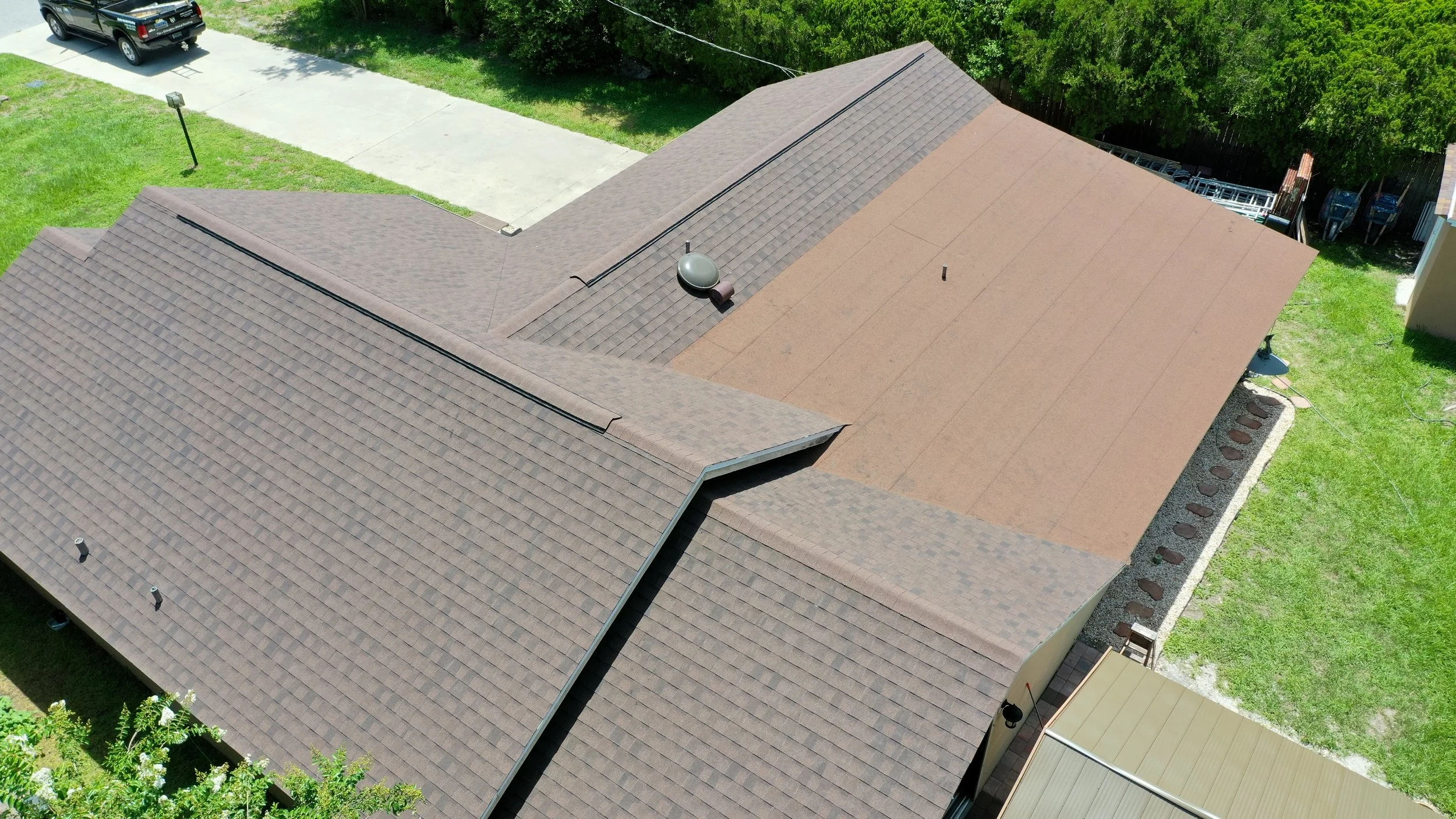 Aerial view of a house roof with brown asphalt shingles and a section of flat brown roofing, surrounded by green grass and a driveway with a pickup truck.