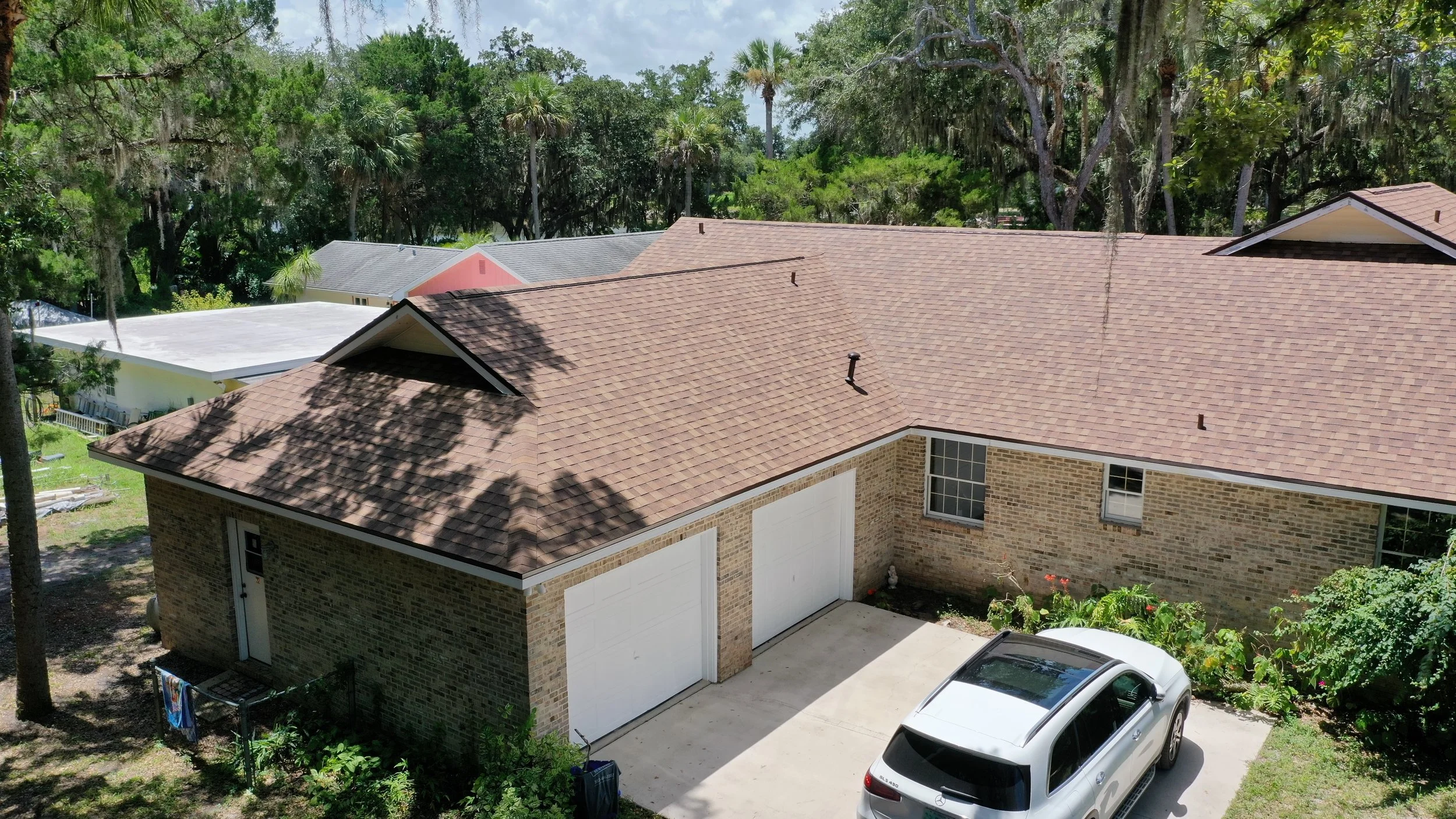 An aerial view of a brick house with a brown tiled roof, a driveway with a silver car, and a backyard with green plants and trees.
