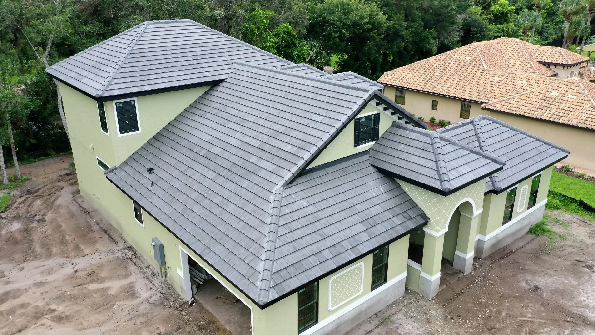 A newly constructed two-story house with light green walls and a grey tiled roof, surrounded by some dirt and grass, with neighboring houses and trees in the background.