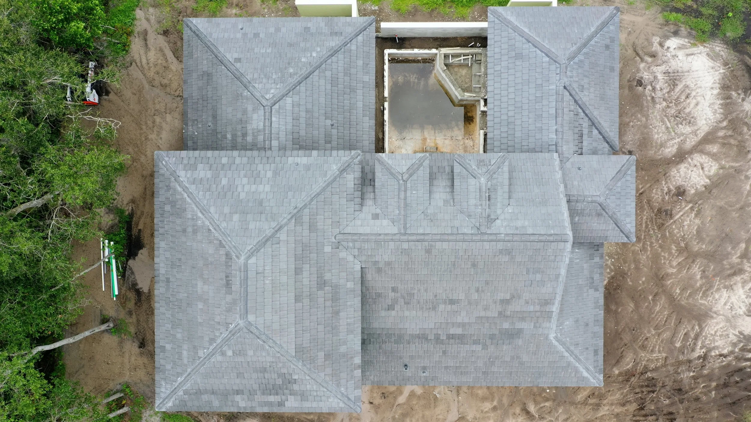An aerial view of a house with grey shingle roof sections, surrounded by partially excavated ground and green trees.