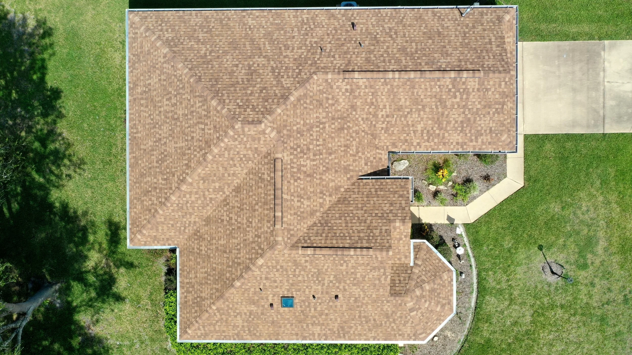Aerial view of a house with a brown shingle roof, surrounded by green grass, trees, a concrete driveway, and a landscaped front yard.