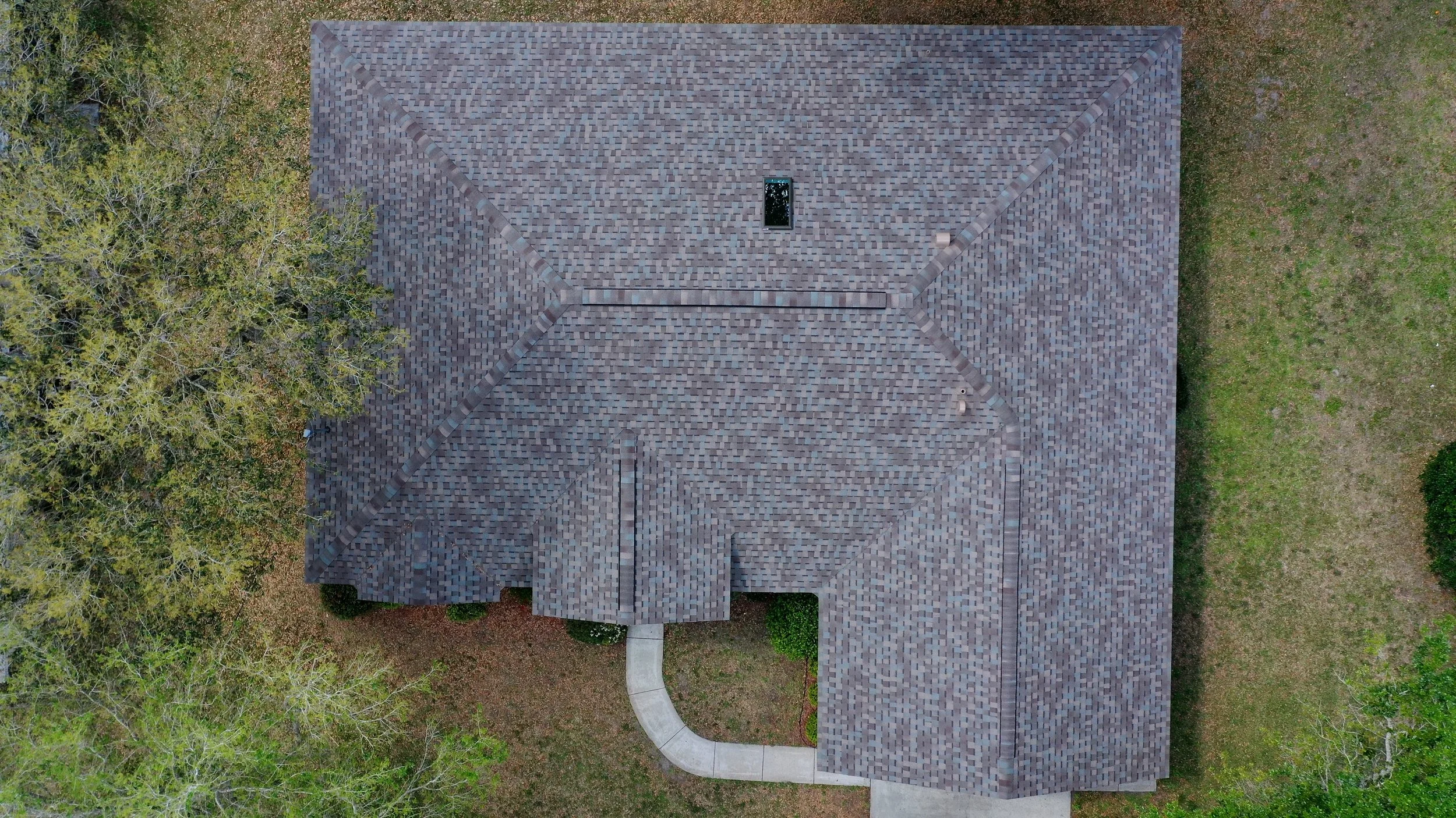 An aerial view of a house with a gray shingled roof, a curved walkway leading to the front door, and trees surrounding the property.