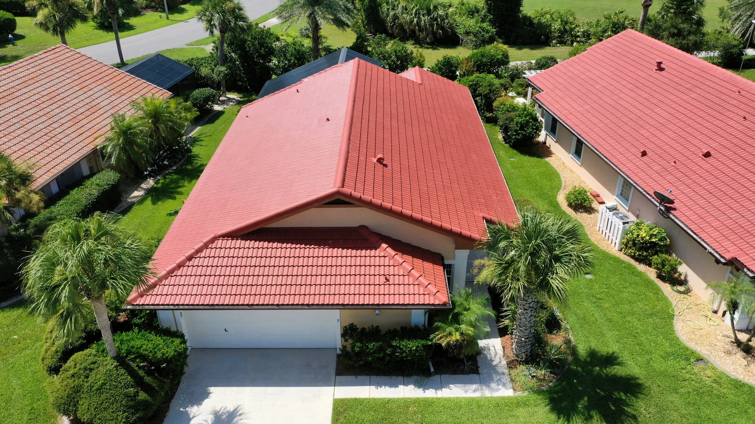 Aerial view of a house with a red-tiled roof, surrounded by green lawns, palm trees, and neighboring houses with similar red roofs.