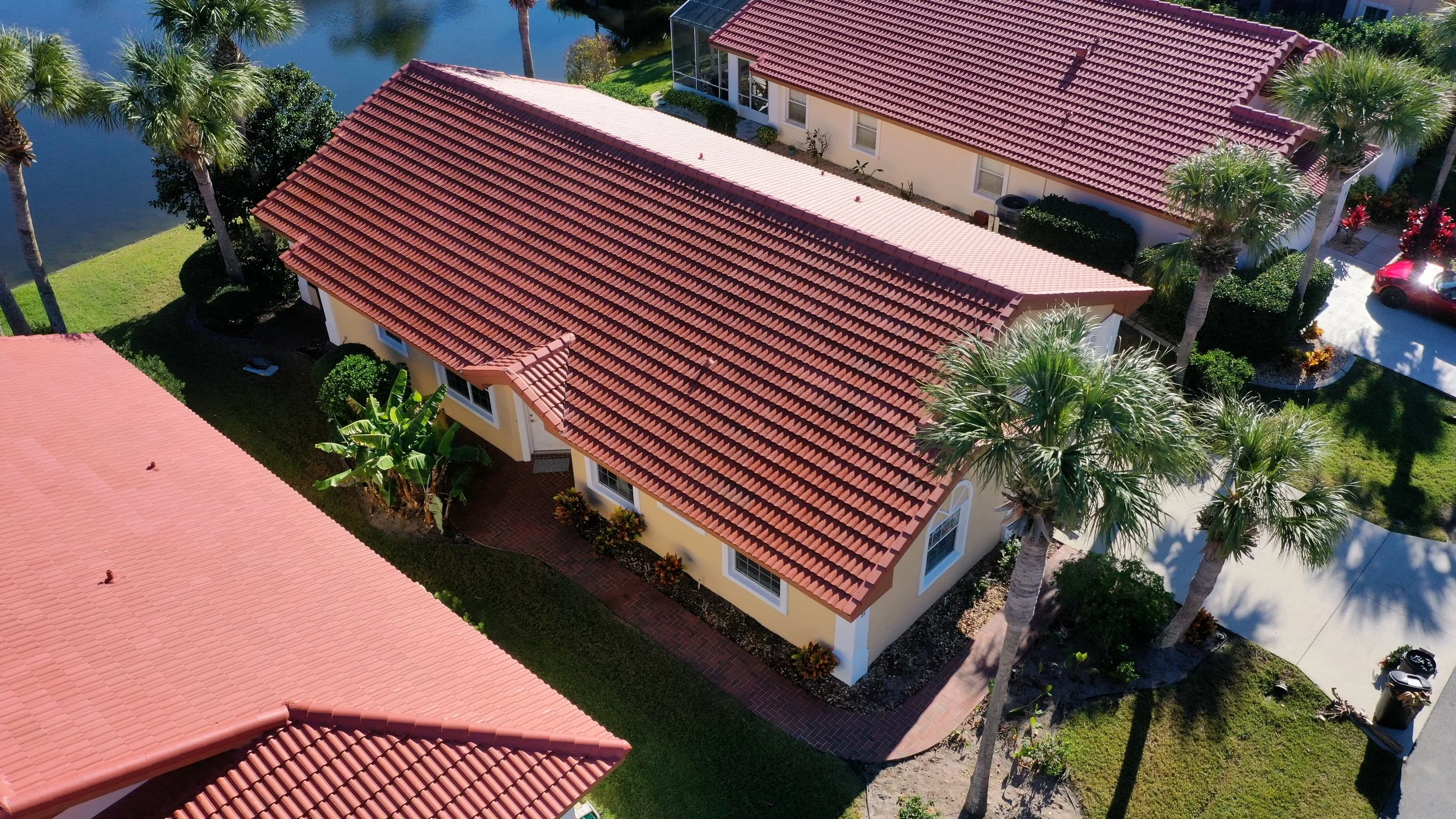 Aerial view of a house with a red tile roof, surrounded by palm trees, a water pond, landscaped yard with a sidewalk, and a parked red car.
