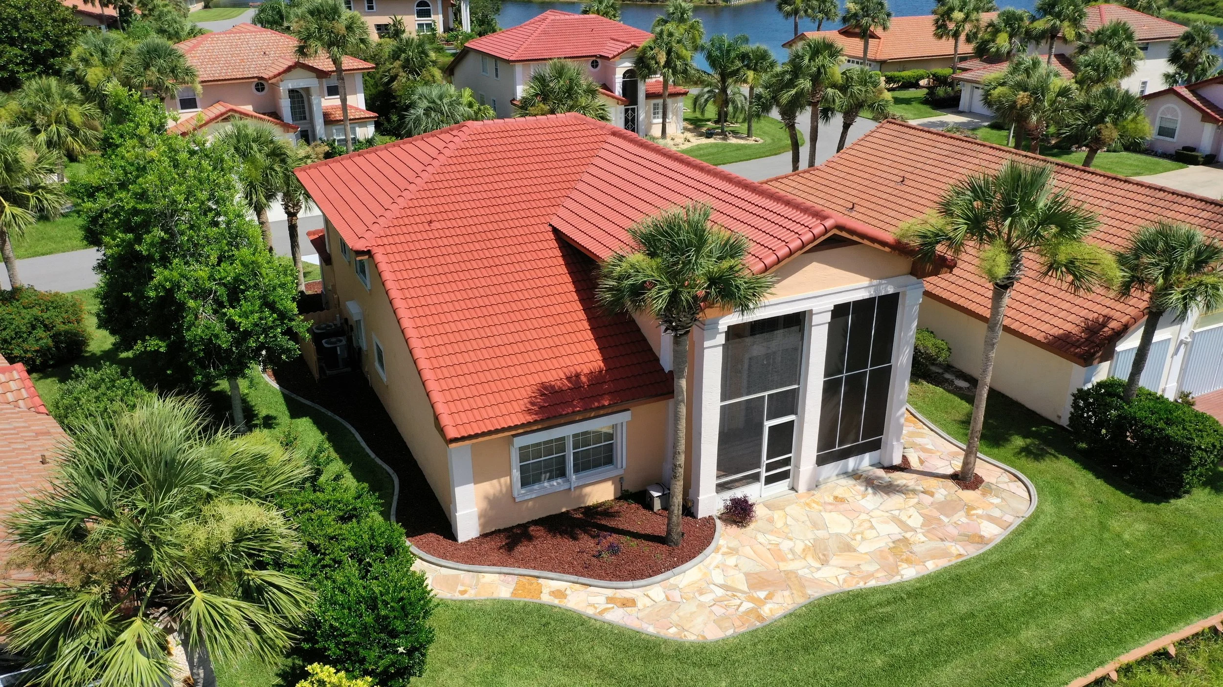 Aerial view of a house with a red tile roof, large front screened porch, and well-maintained lawn, surrounded by palm trees in a neighborhood with other similar houses and a water body in the background.