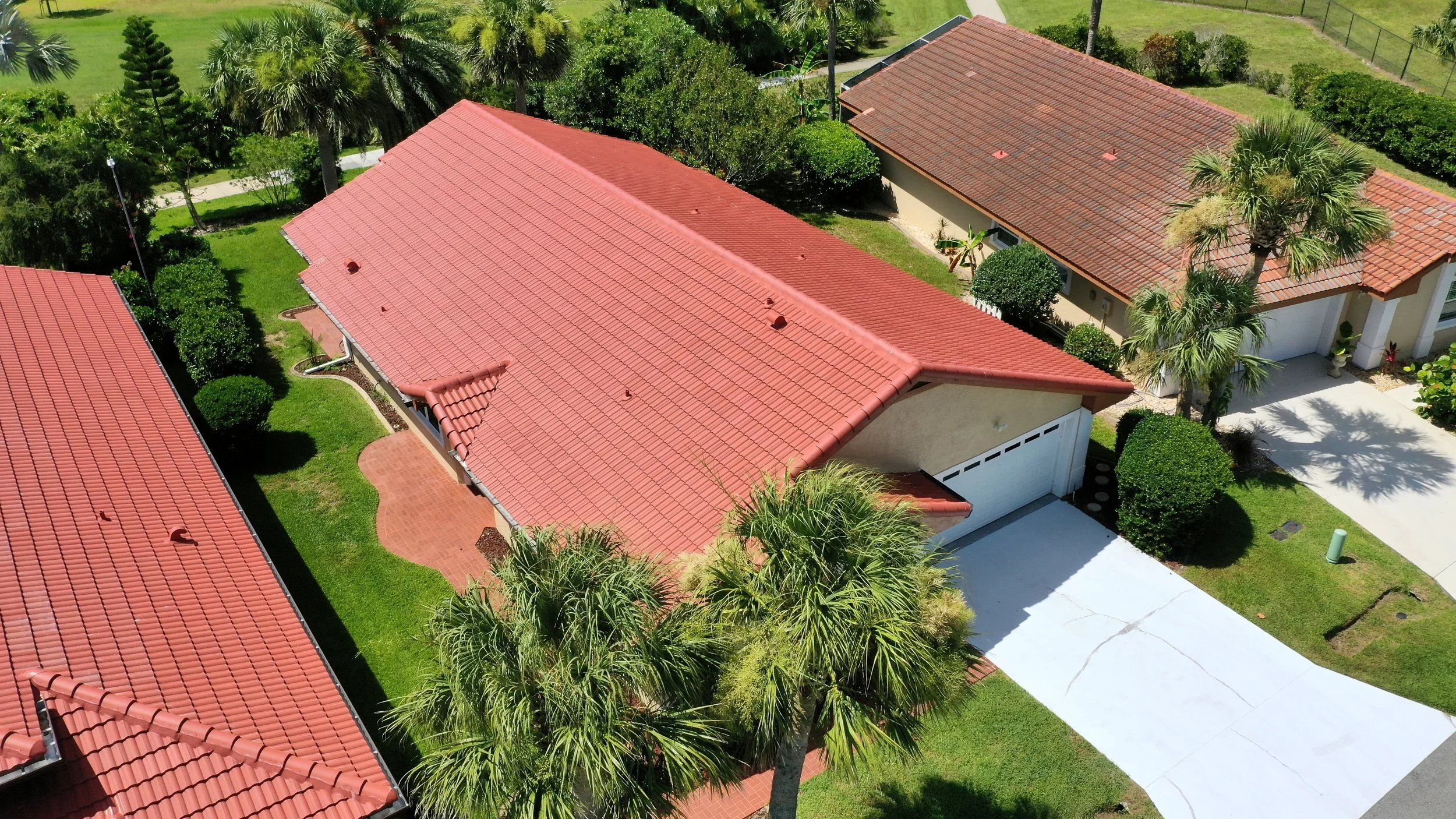 Aerial view of a house with a red-tiled roof and a white garage door, surrounded by a manicured lawn, palm trees, and neighboring houses with similar red-tiled roofs in a suburban neighborhood.
