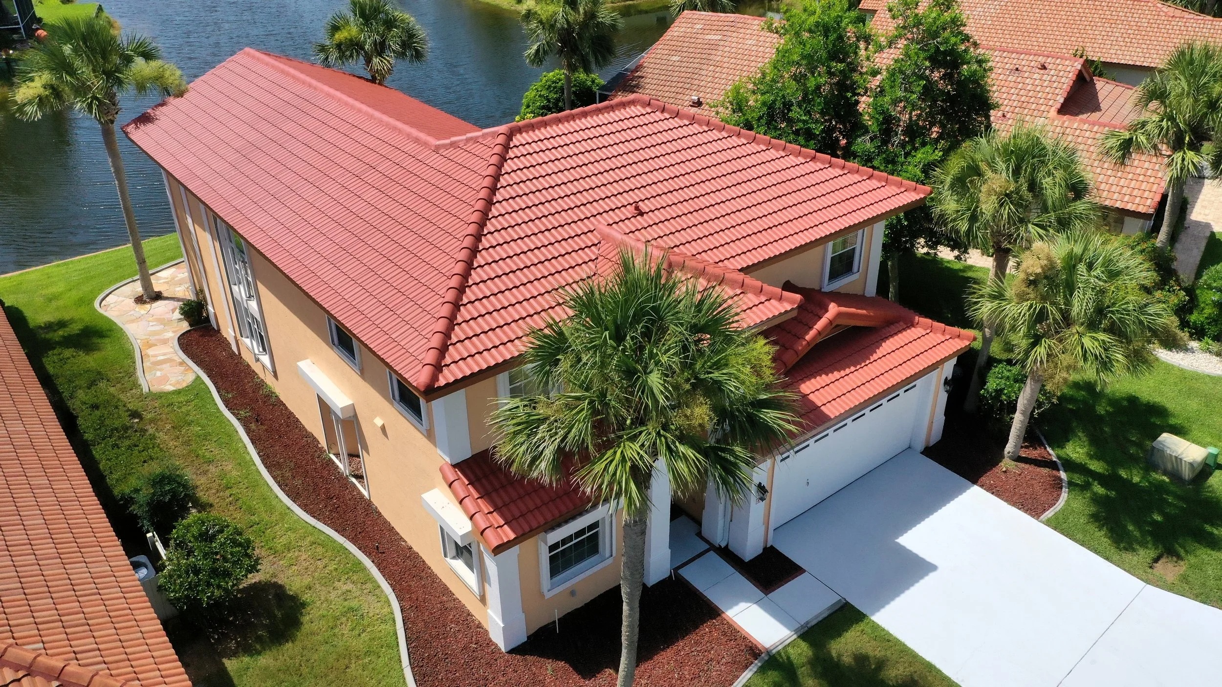 Aerial view of a two-story house with a red tile roof, beige exterior walls, and a white garage door, surrounded by green lawn, palm trees, and neighboring houses with similar roofs.