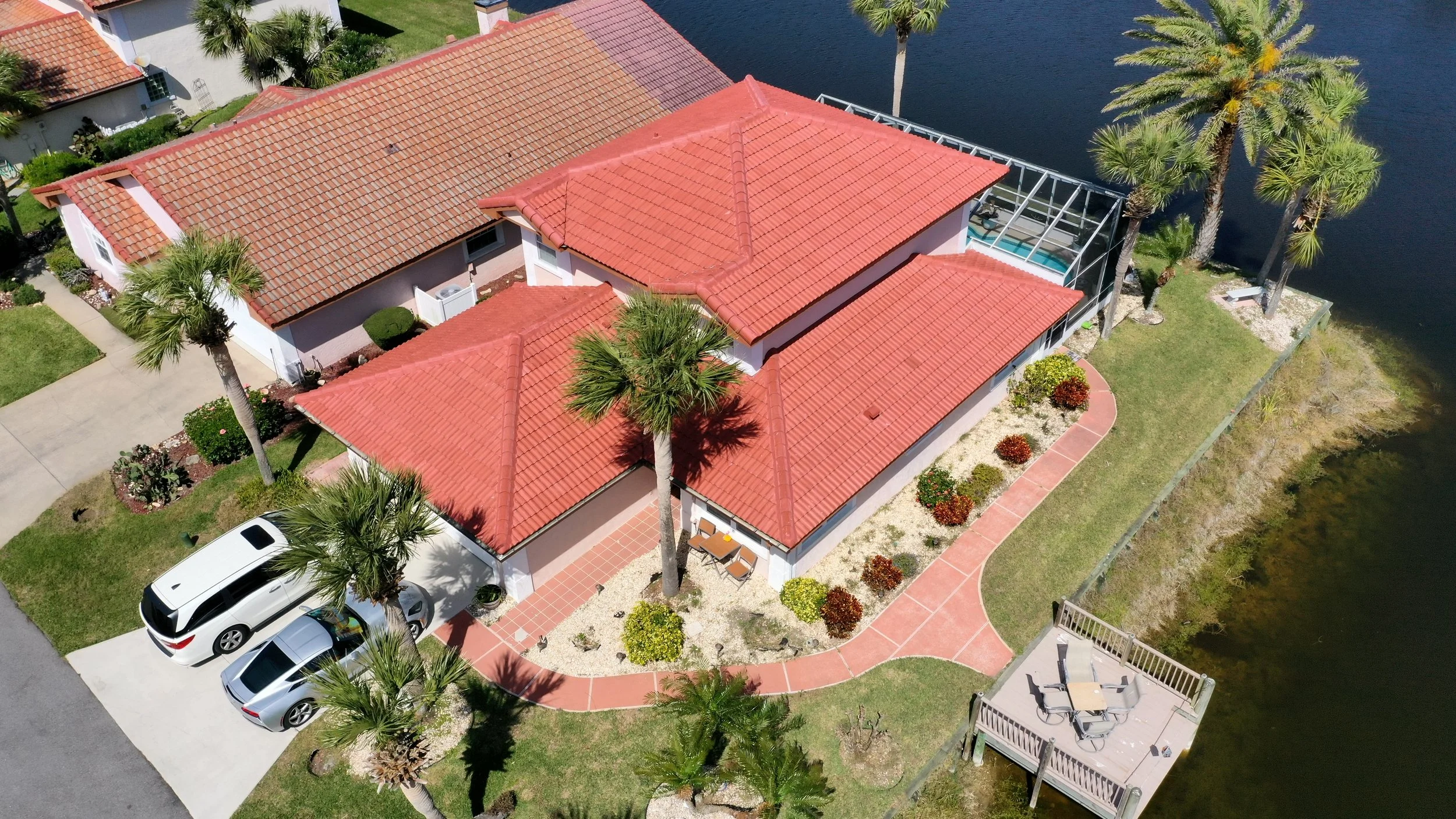 Aerial view of a house with a red tile roof located by a body of water, surrounded by palm trees, a landscaped yard with bushes, a driveway with two parked cars, and a deck with outdoor furniture.