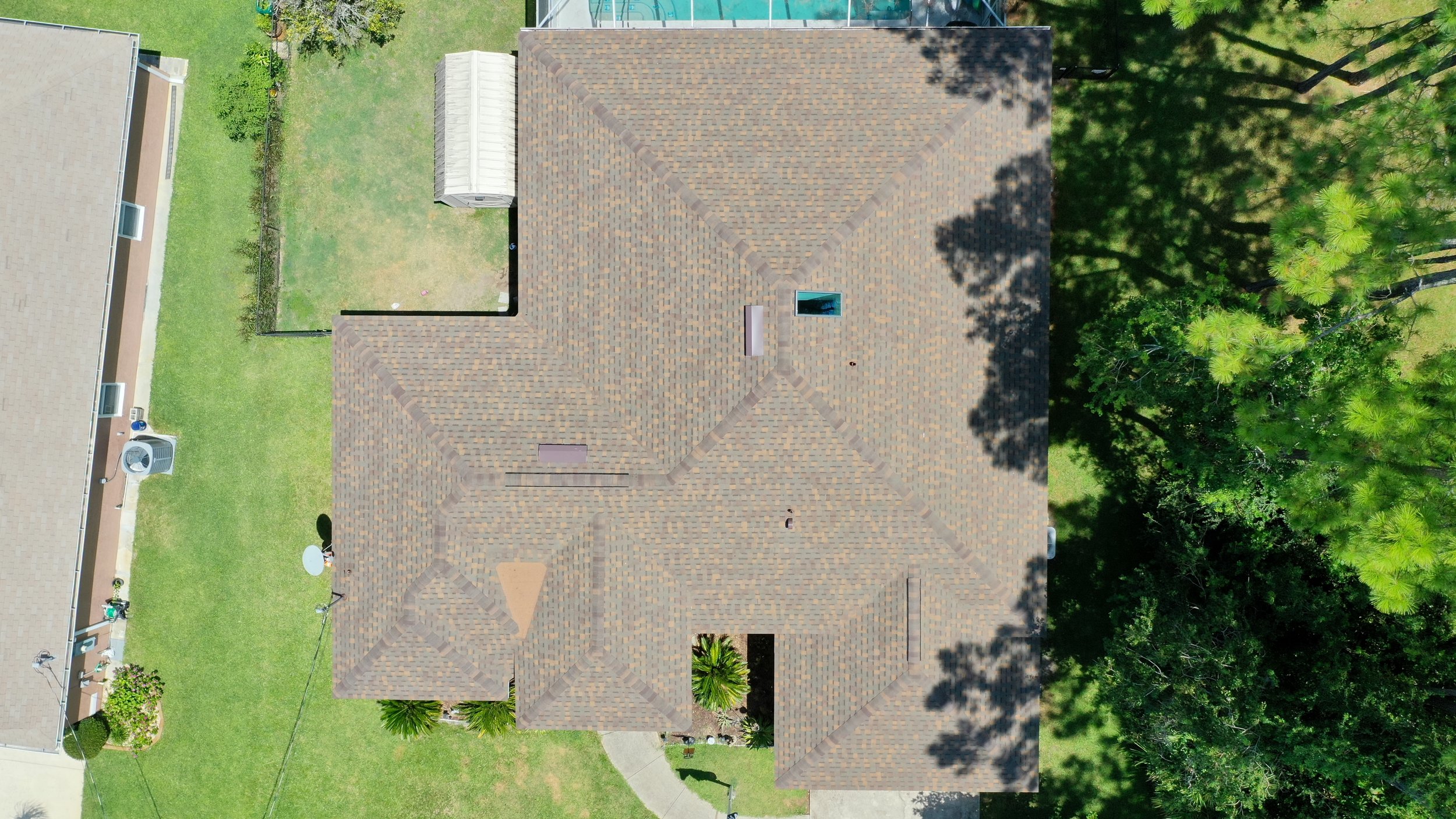 Top view of a house with a brown shingle roof, surrounded by green trees and a lawn, with neighboring houses visible on the left.
