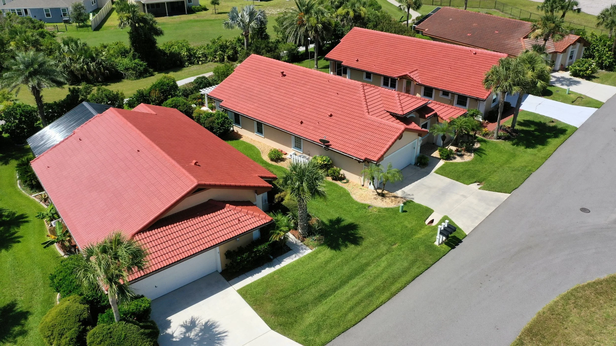 Aerial view of a suburban neighborhood with three houses, each with red tile roofs and surrounded by green lawns, trees including palm trees, and driveways. The neighborhood appears sunny and well-maintained.