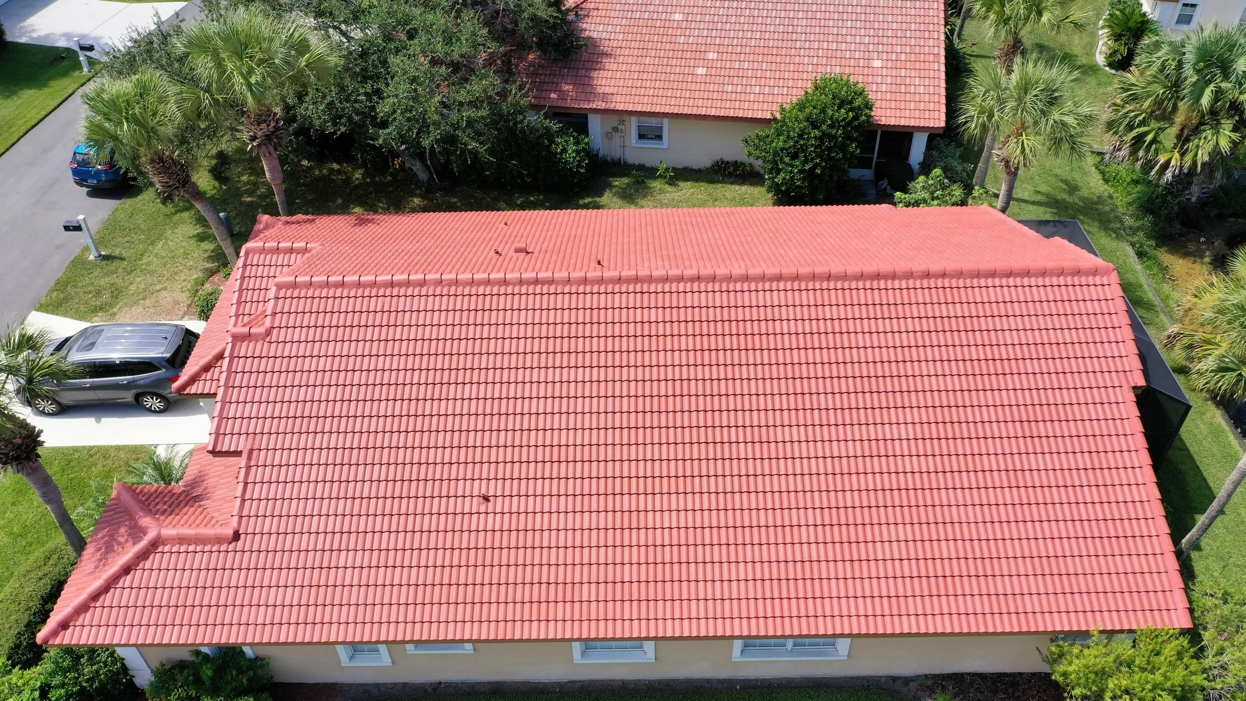 Aerial view of a house with a red-tiled roof, surrounded by palm trees and green lawns, with parked cars and neighboring houses.