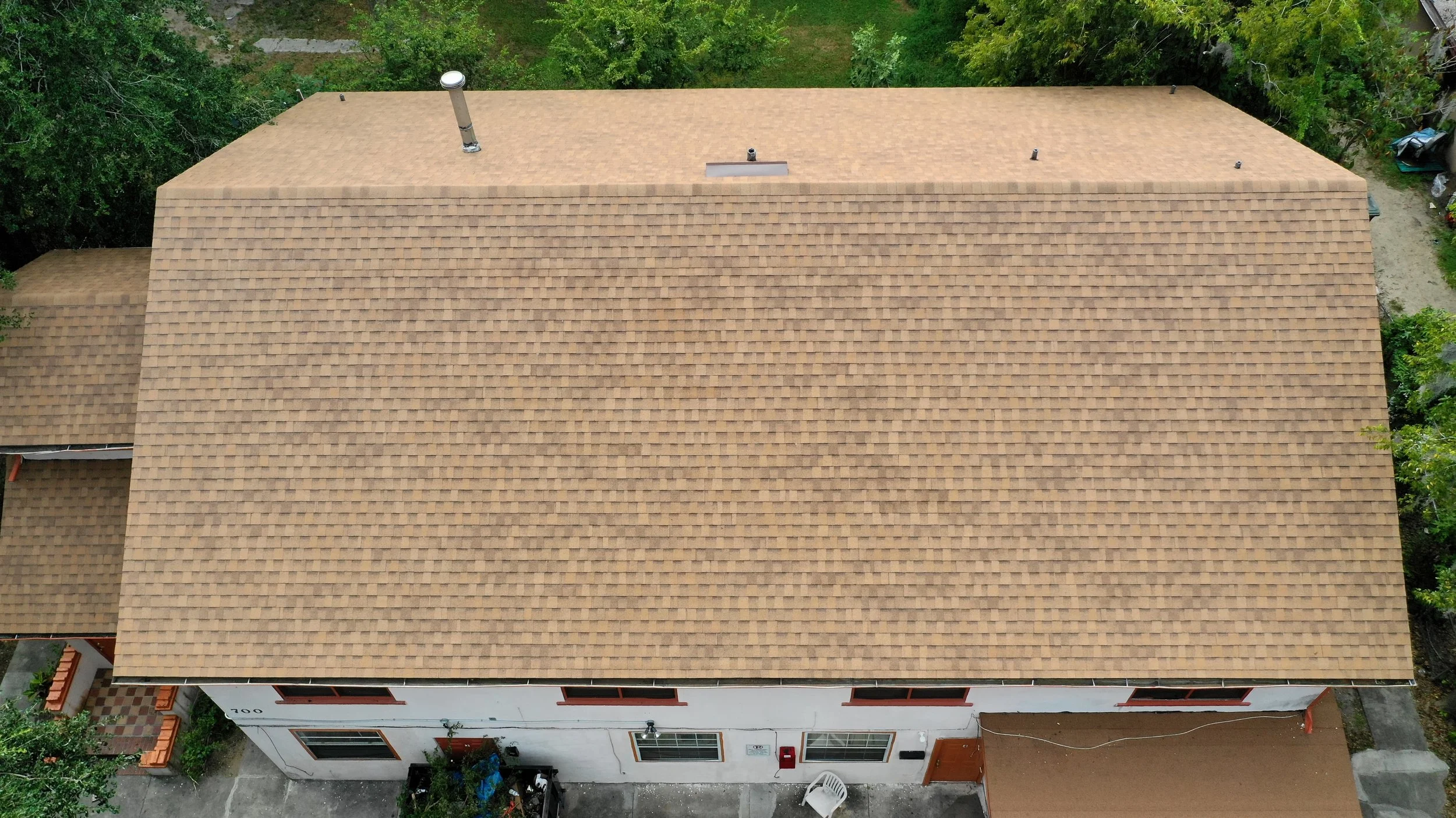Aerial view of a house with a large, tan shingle roof surrounded by green trees and neighboring buildings.
