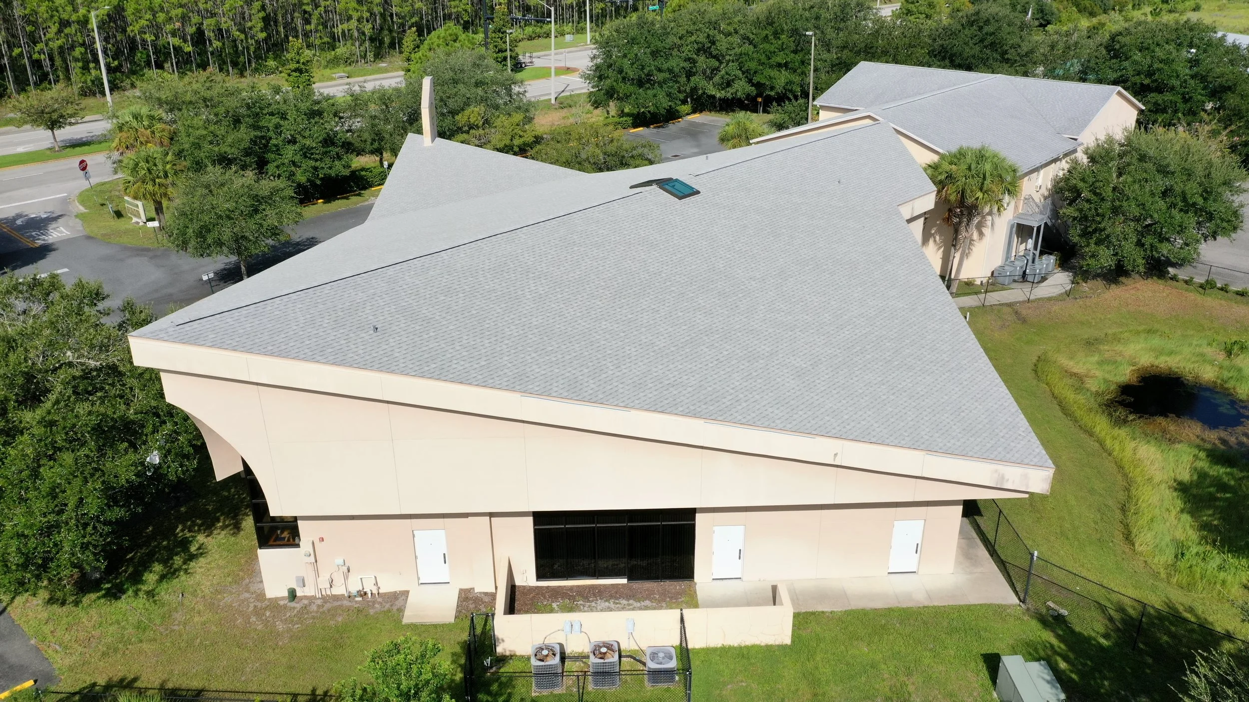 Aerial view of a modern building with a unique angular roof, surrounded by green trees, grass, and a small pond.