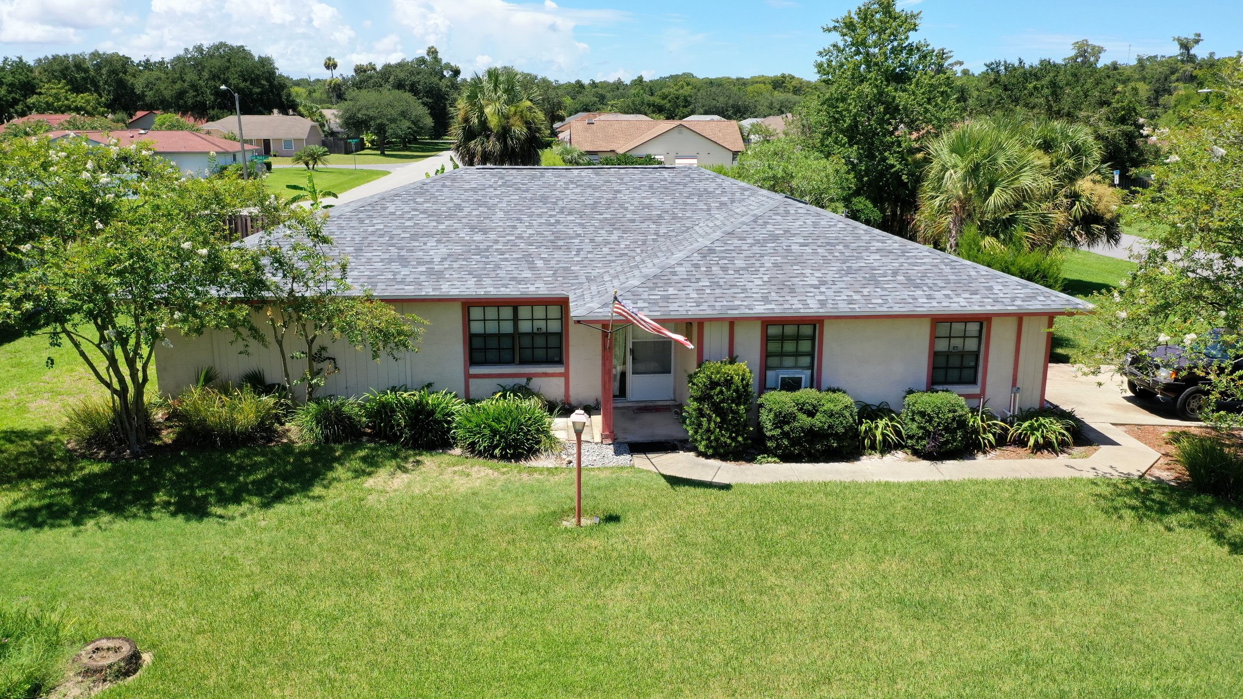 A single-story house with a gray shingle roof, white exterior walls with red trim, surrounded by greenery, and a lawn. An American flag hangs near the front door. A black vehicle is parked on the driveway to the right.