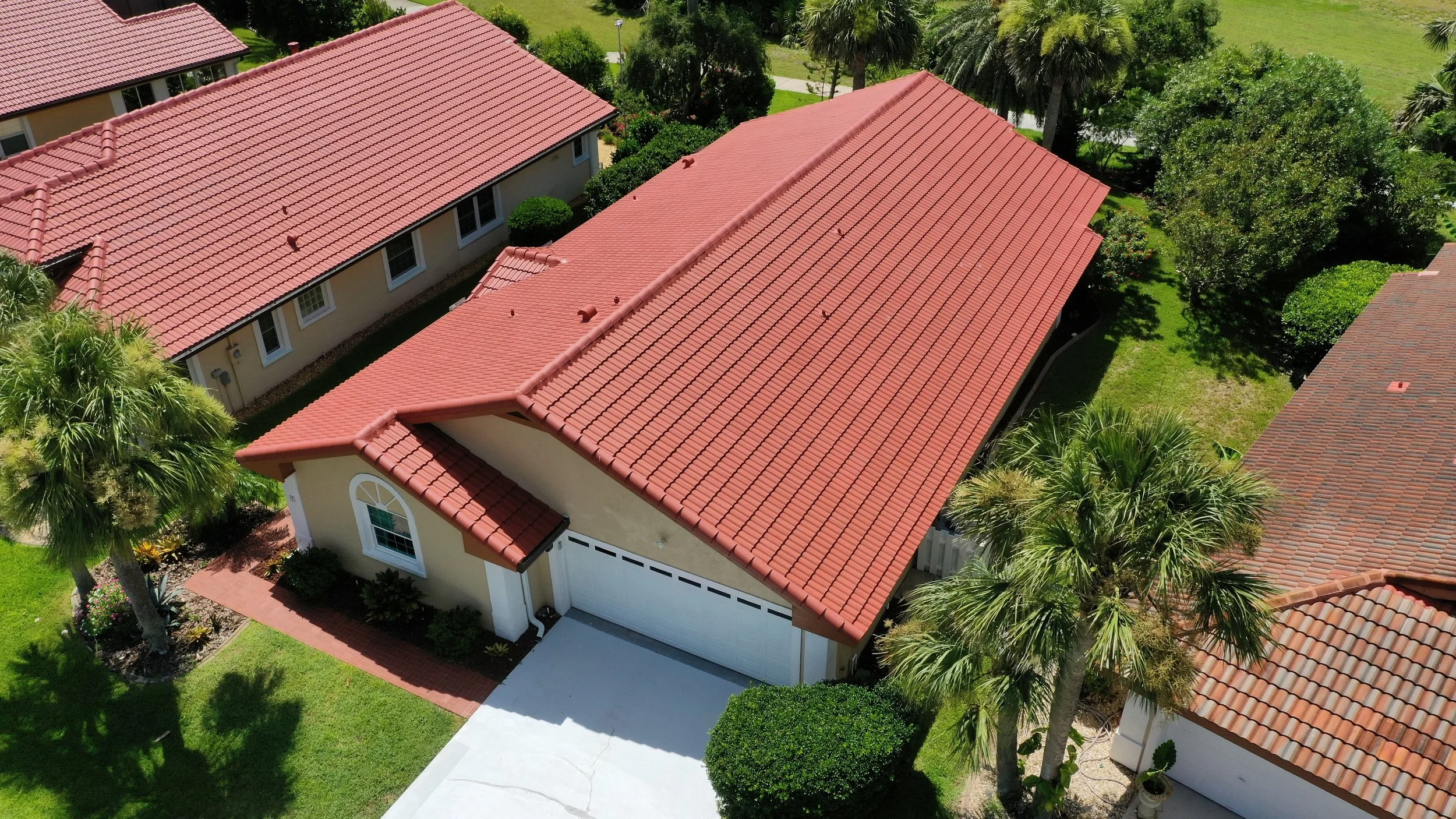 Aerial view of a residential neighborhood showing houses with red tile roofs, green lawns, and trees, including palm trees.