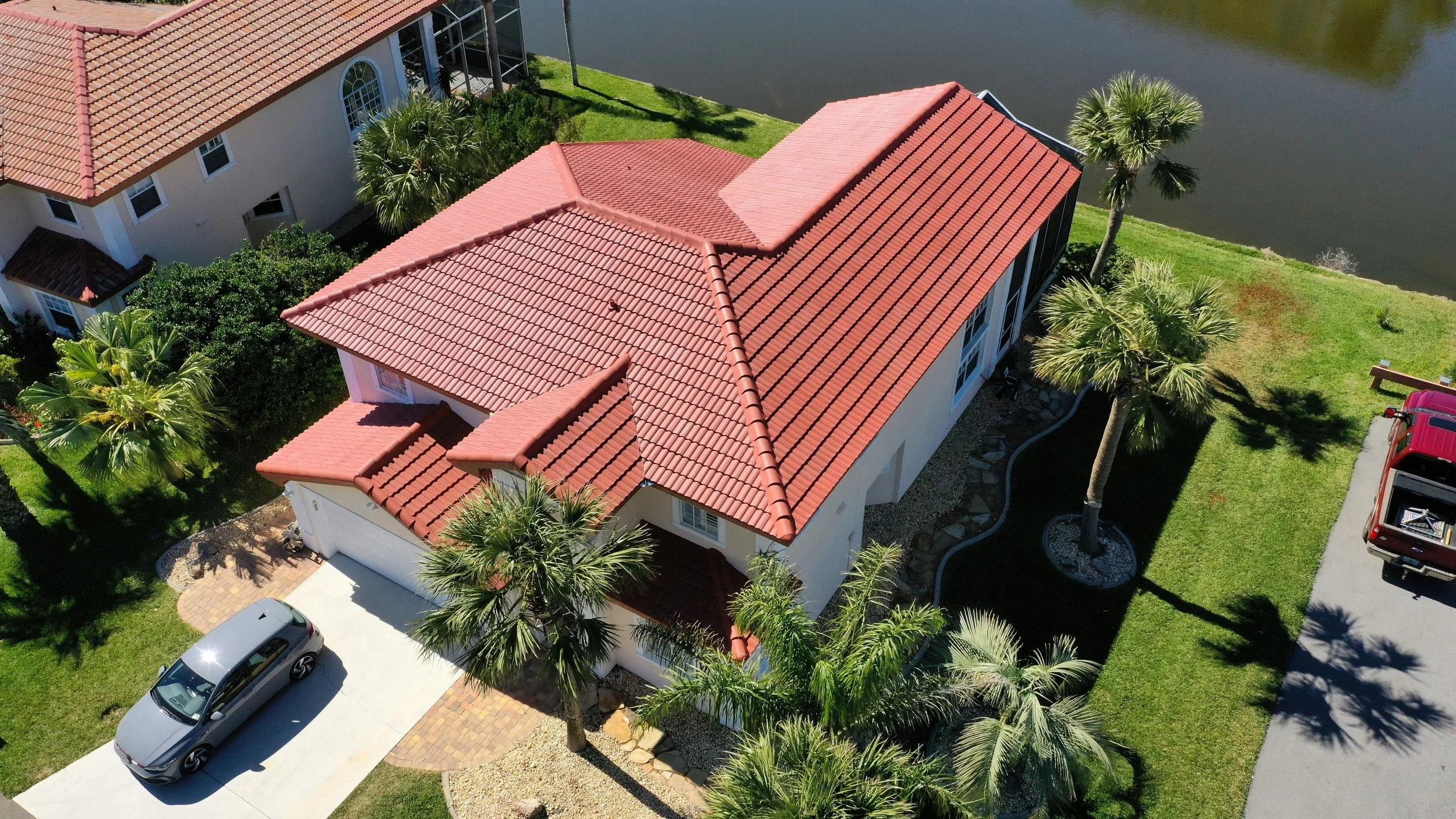 Aerial view of a house with a red tiled roof, surrounded by palm trees, next to a water body, with cars parked in the driveway.