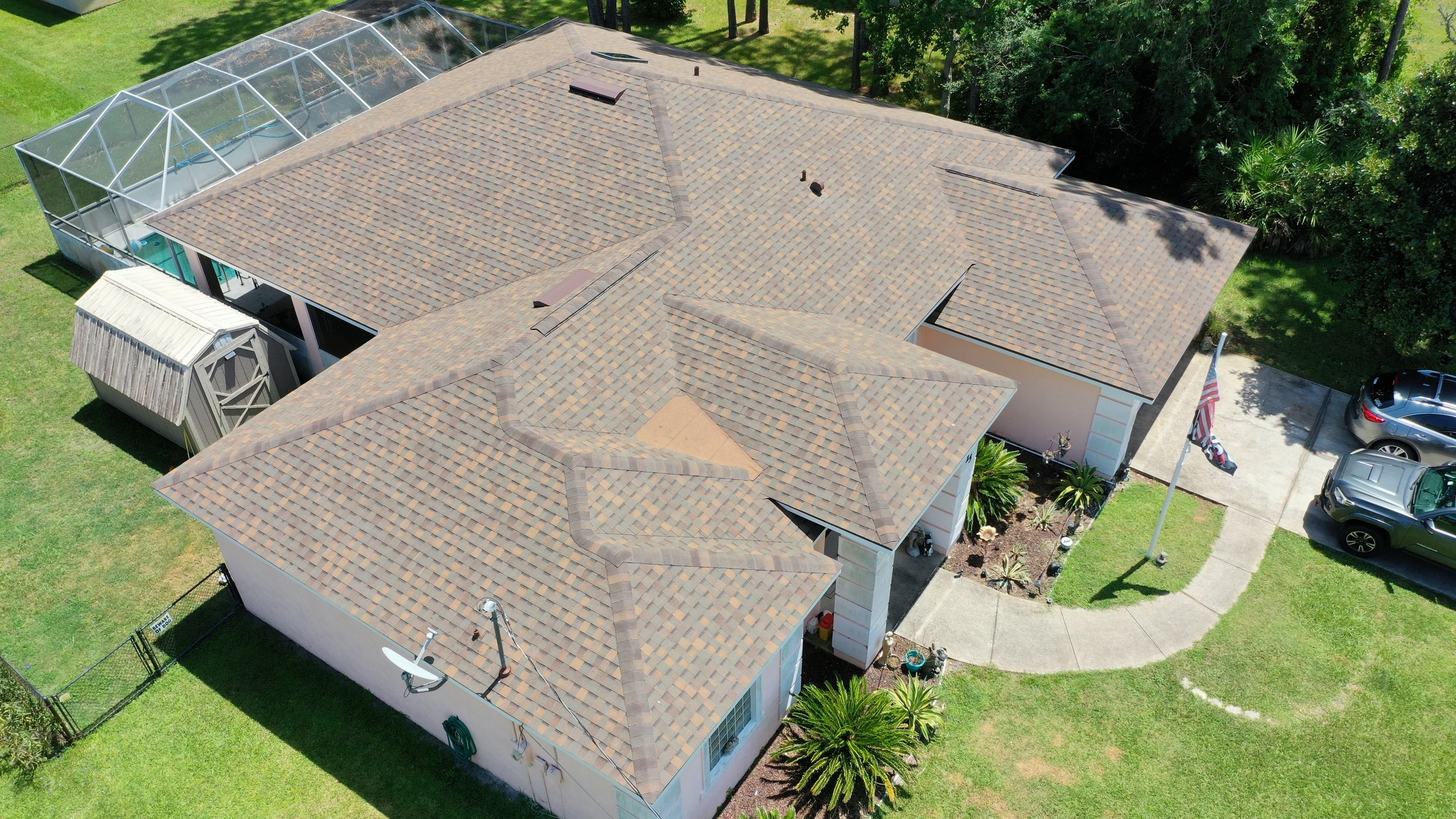 Aerial view of a house with a tiled roof, a small backyard with a lawn, outside patio, and a screened-in pool enclosure, surrounded by trees and parked cars.