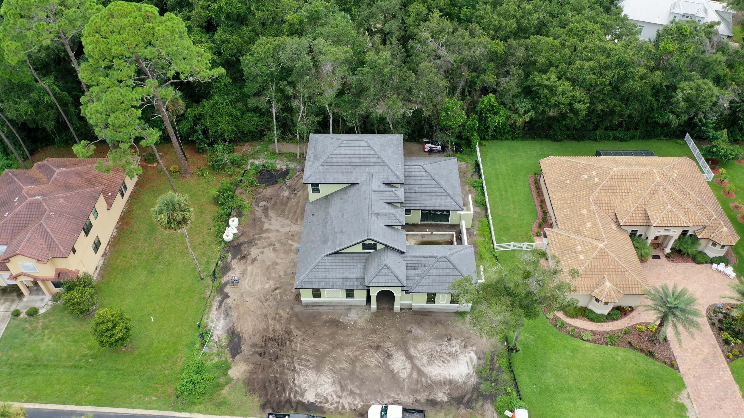 An aerial view of a house under construction with a gray roof, surrounded by neighboring completed houses with tiled roofs, green lawns, and trees.