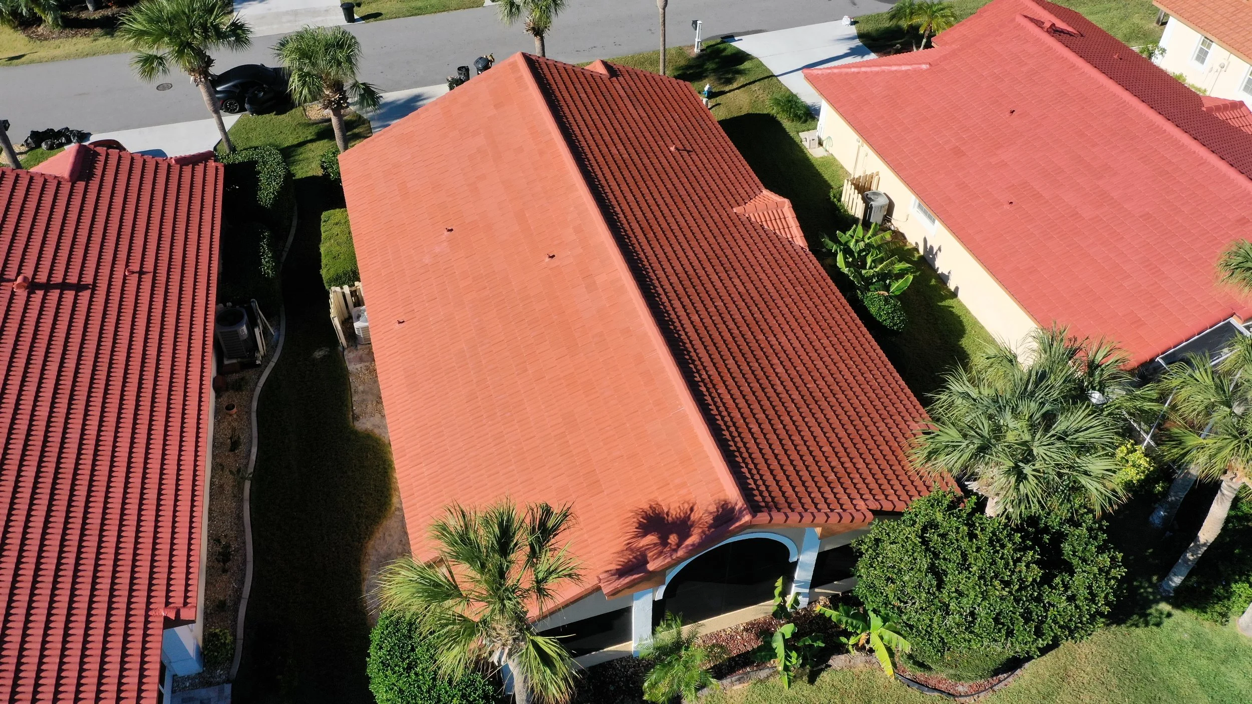 Aerial view of several houses with red-tiled roofs, surrounded by palm trees, green bushes, and a lawn in a suburban neighborhood.